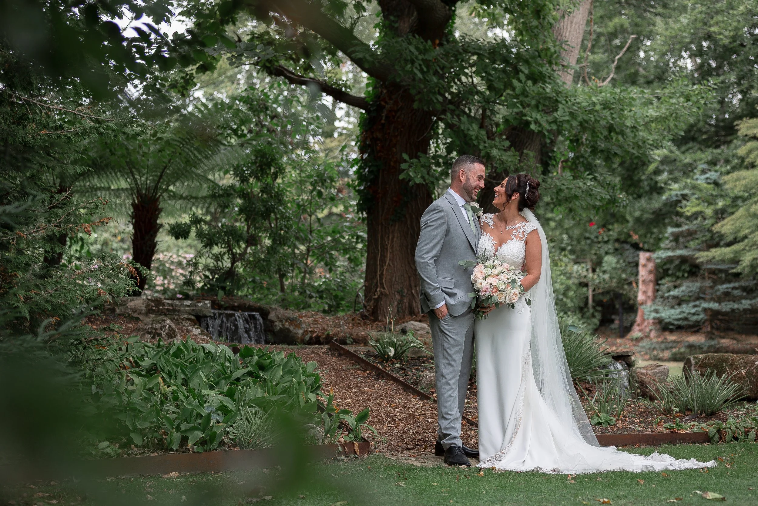 A bride and groom stand close together outdoors by a large tree, smiling and gazing into each other's eyes, surrounded by lush green foliage.