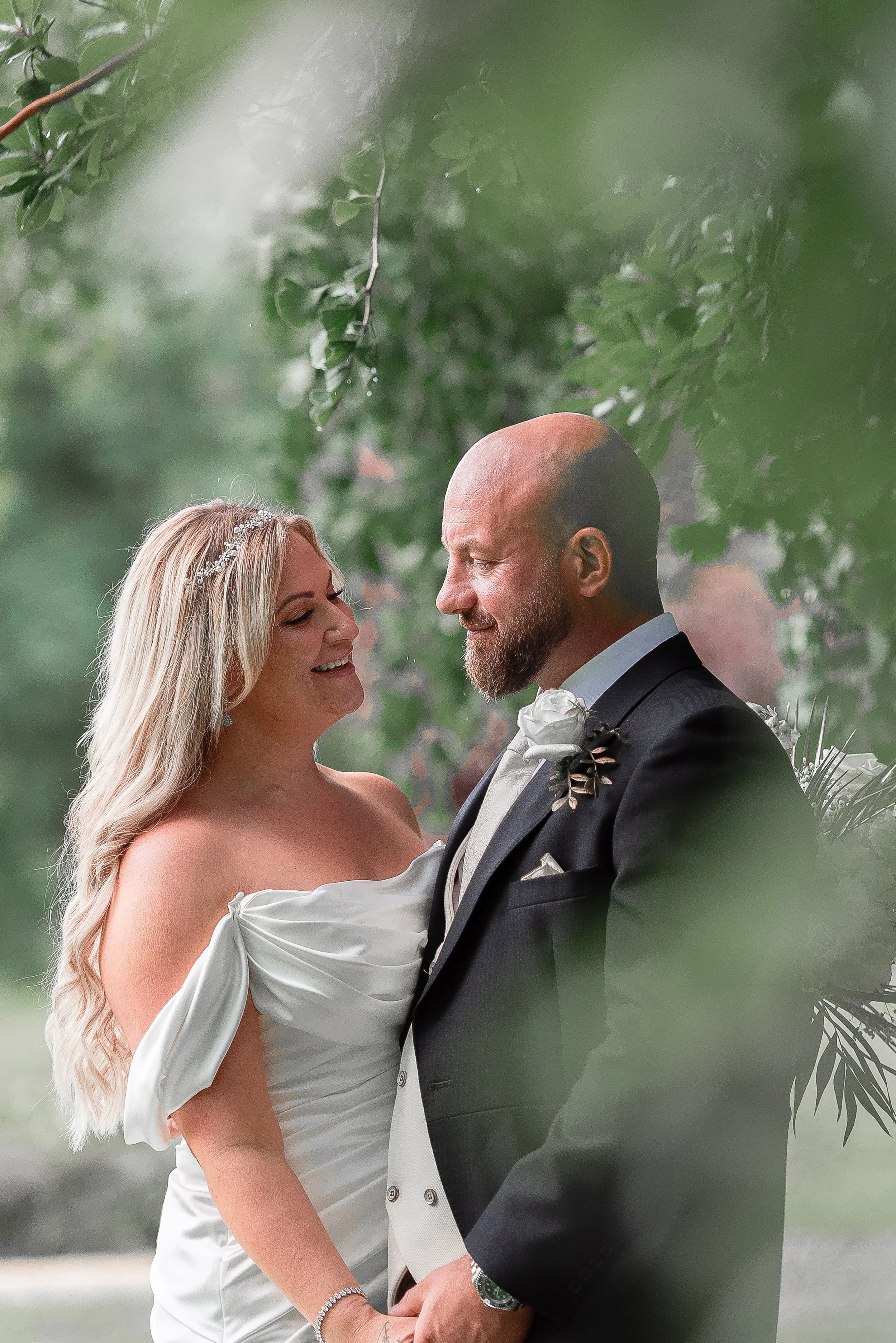 A bride and groom are smiling and looking into each other's eyes at their wedding ceremony, holding hands. The bride has long blonde hair, a pearl bracelet, and a strapless white dress with puffed sleeves. The groom is bald, has a beard, and is weari