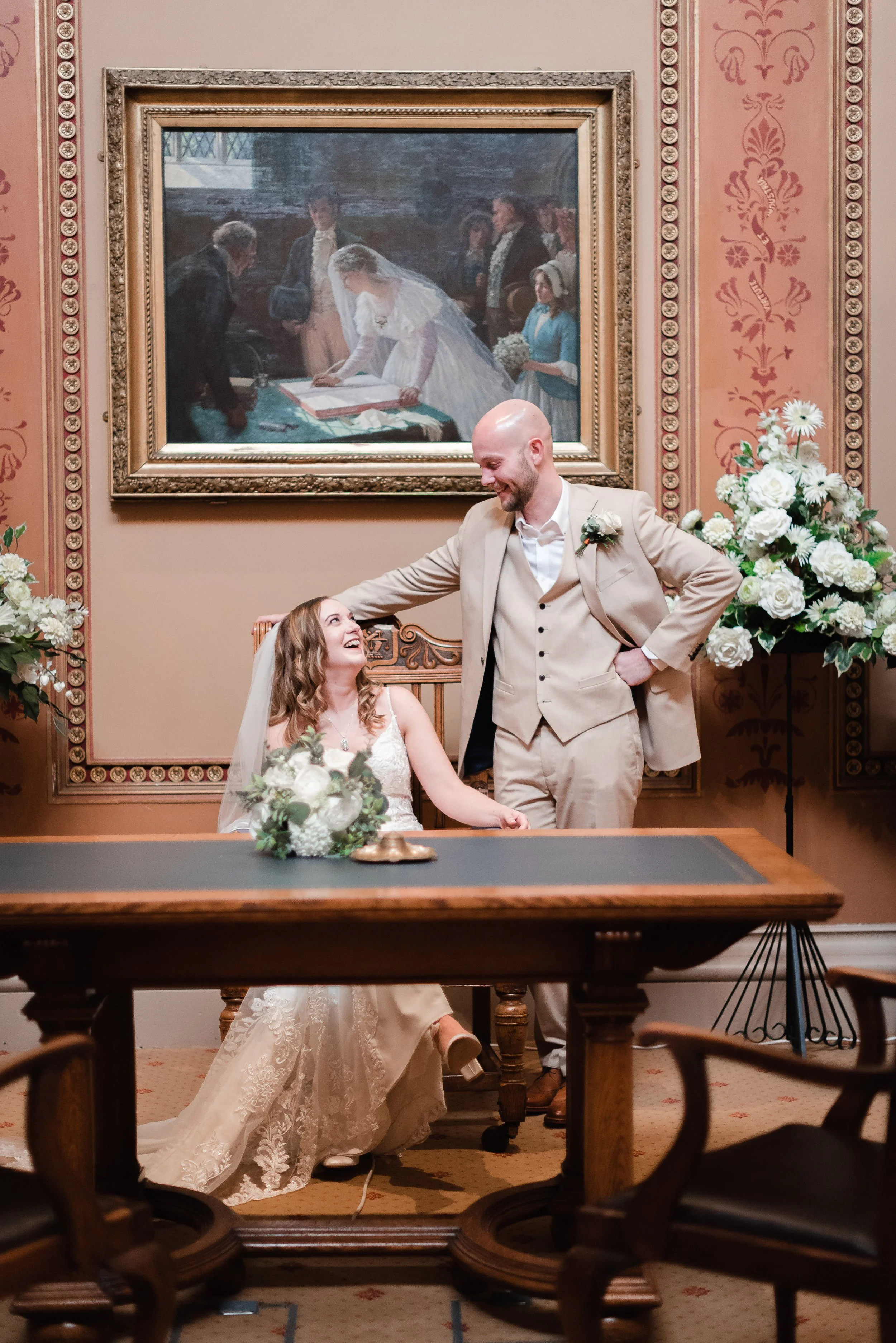 A bride and groom at a wedding signing documents, with the groom standing and the bride seated and holding a bouquet, in a room with floral wallpaper, large floral arrangements, and a large framed painting of a wedding scene hanging on the wall behin