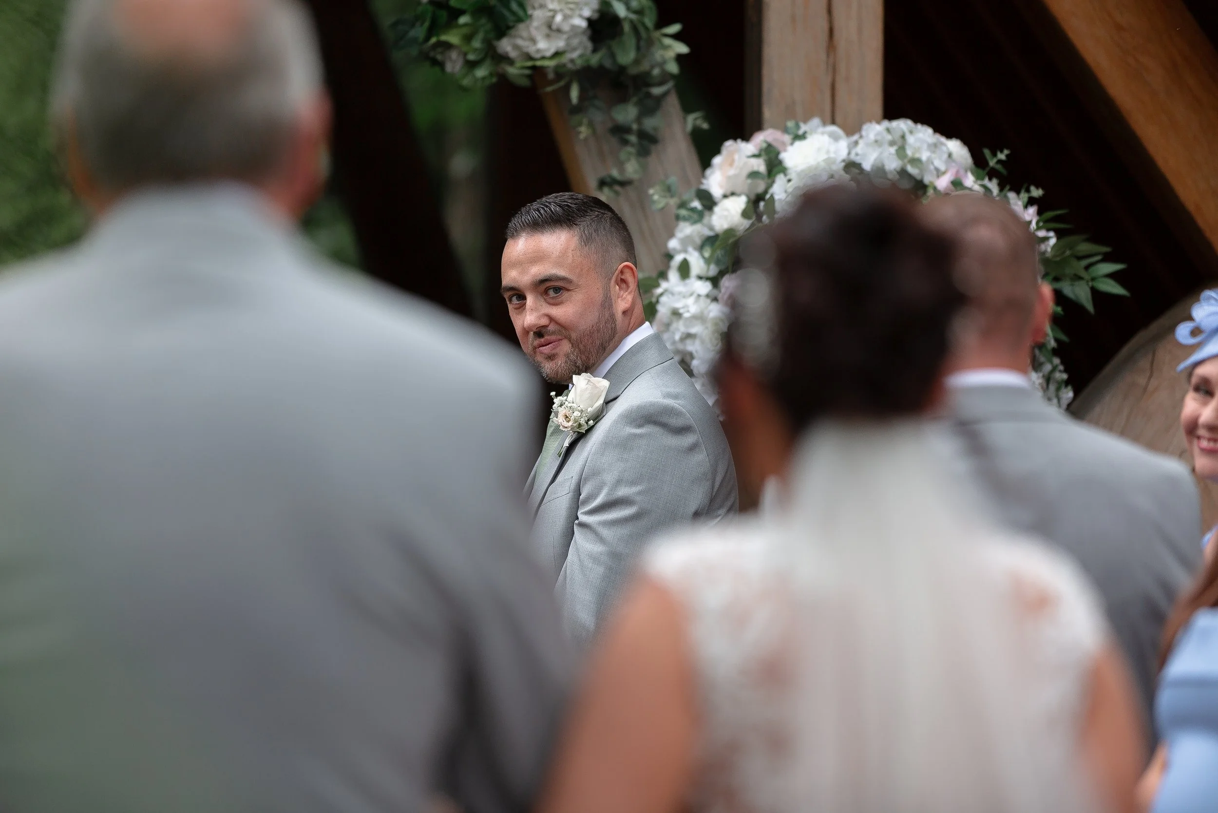Man in a gray suit looking over his shoulder during a wedding ceremony with floral decorations in the background.