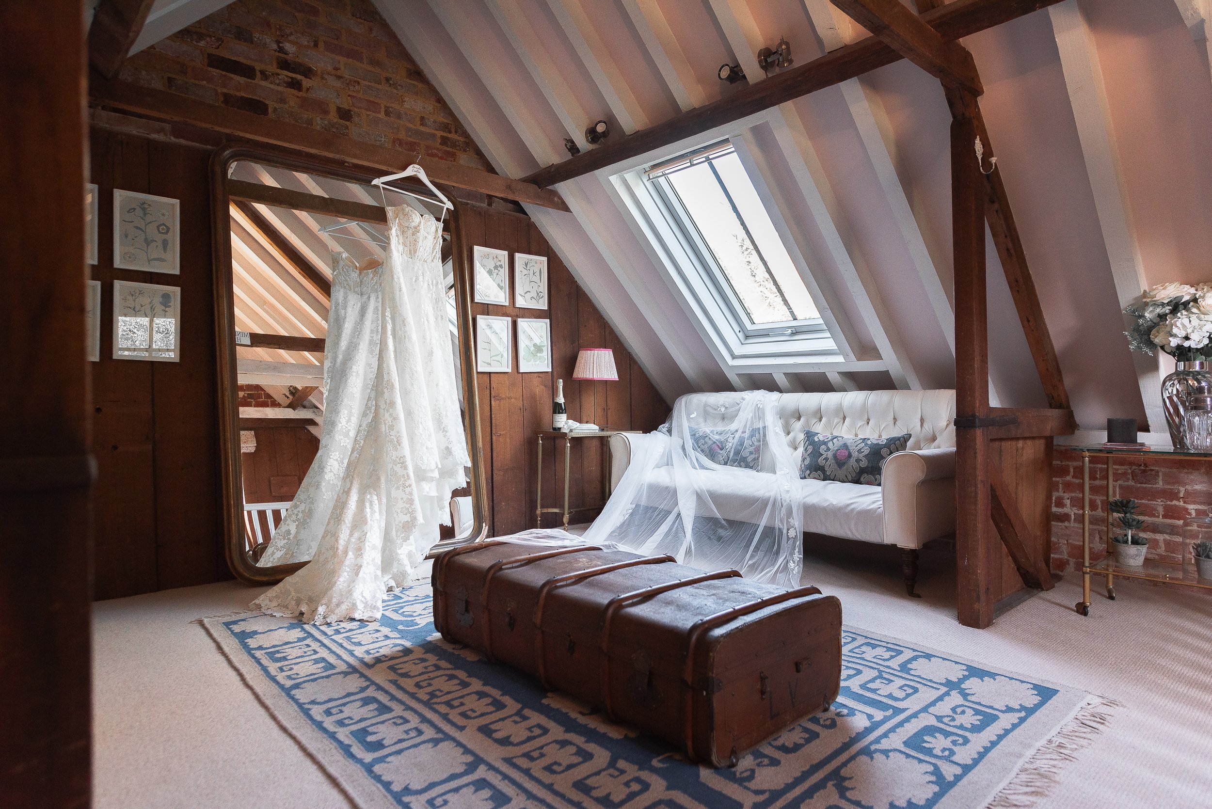 A cozy attic room with a sloped ceiling featuring a skylight window, a vintage white sofa with pillows, a wooden dresser, and a dress hanging on a mirror door. The room has wooden walls and exposed brick accents, and a blue patterned rug on the floor