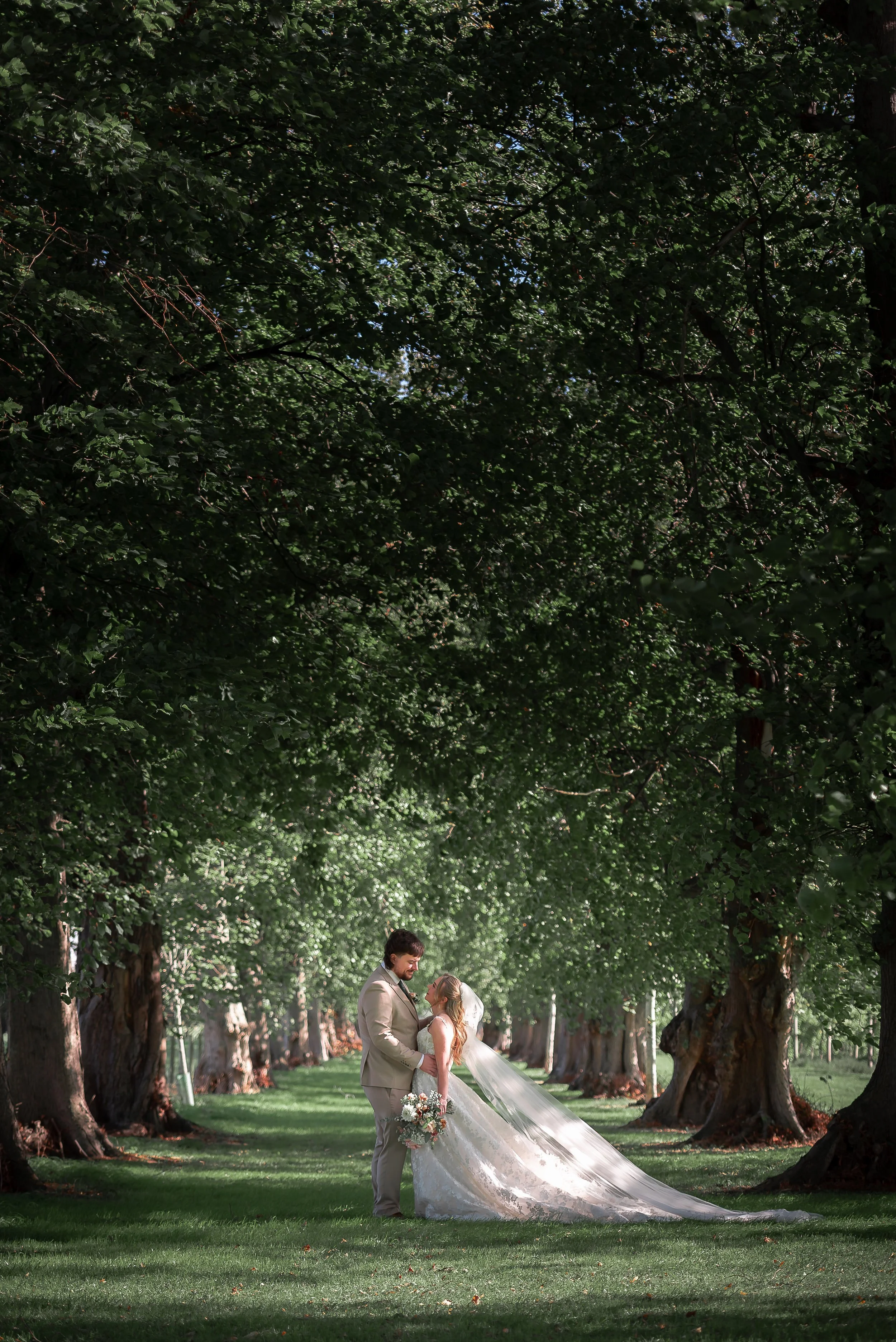 A bride and groom standing close together, holding hands, in a tree-lined park during daytime, with the bride wearing a white wedding dress and holding a bouquet, and the groom wearing a beige suit and tie, under a canopy of large green trees.