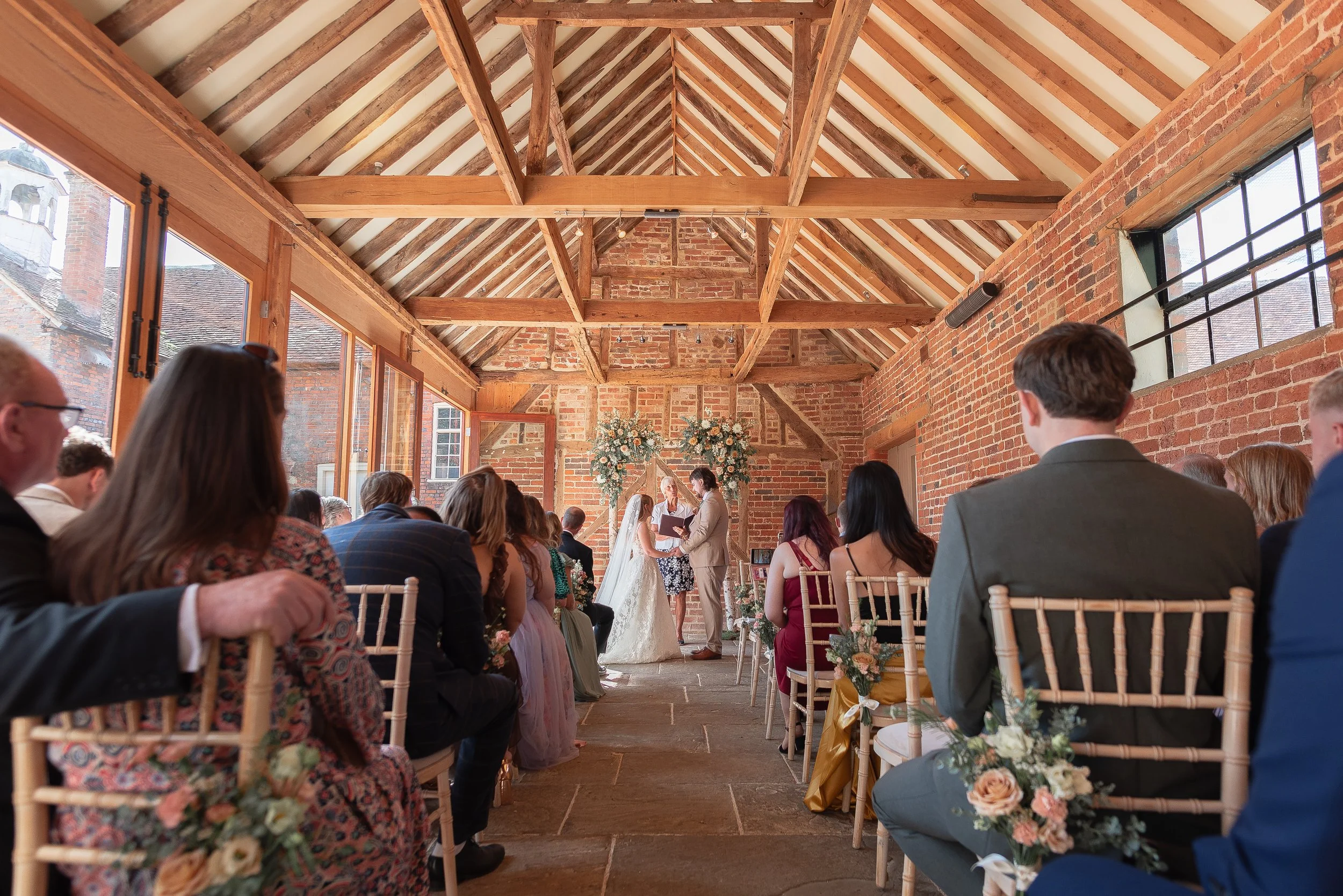 Indoor wedding ceremony in a rustic brick and wood venue with seated guests facing an altar where a couple is exchanging vows, decorated with floral arrangements.
