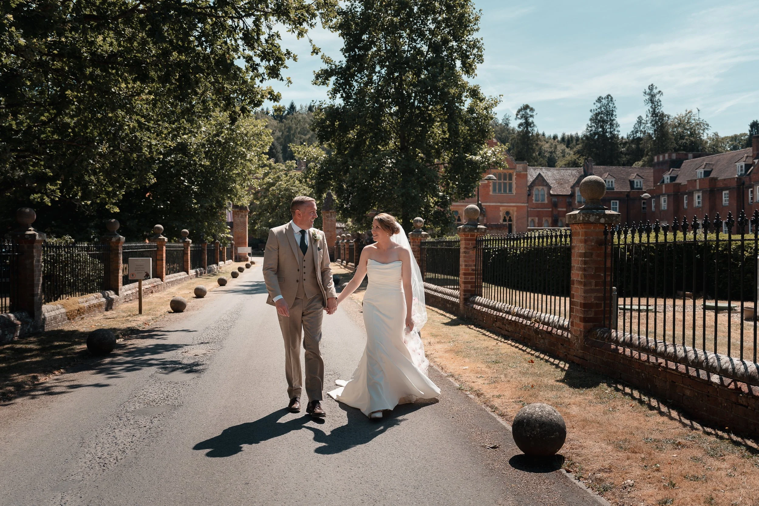 A newlywed couple holding hands and smiling while walking outdoors on a sunny day, with large green trees and historic brick buildings in the background.