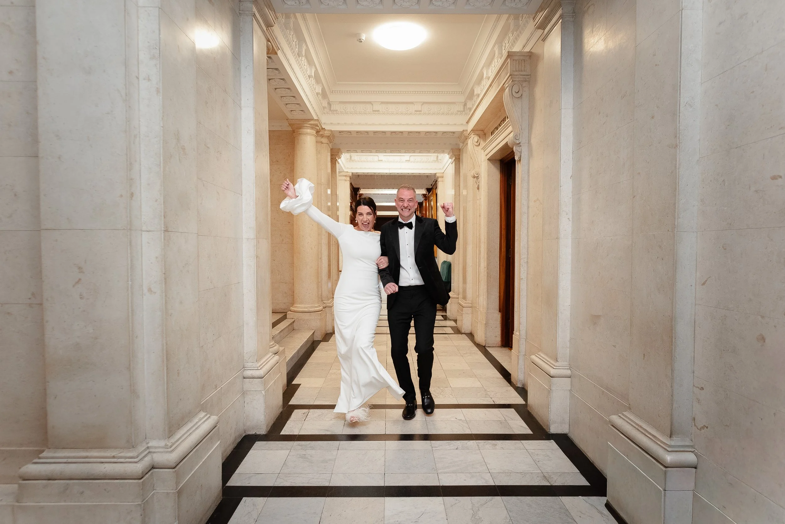 A joyful couple in wedding attire celebrating inside Marylebone Registry Office, with the bride in a white wedding dress and the groom in a black tuxedo, standing in a marble-lined corridor.