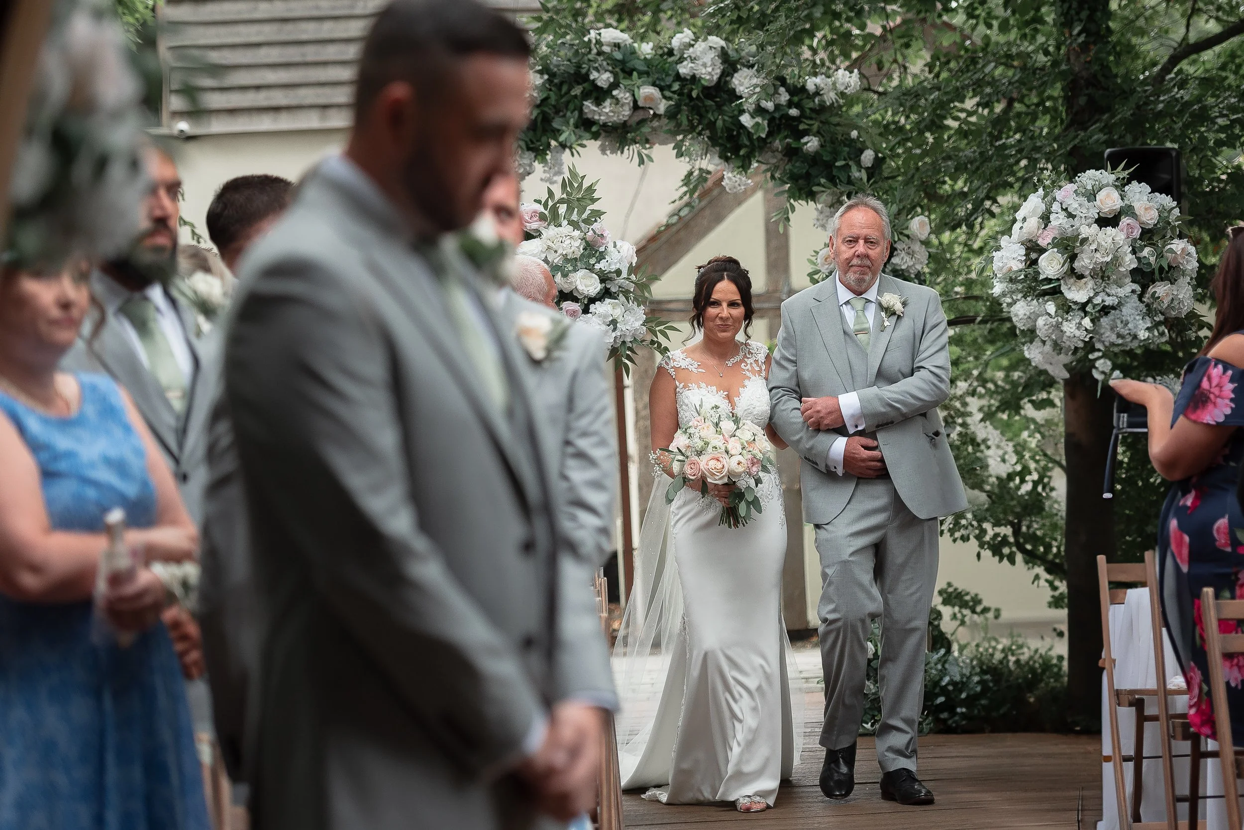 A bride and a man, likely her father, walking down the aisle at an outdoor wedding ceremony, surrounded by floral arrangements and guests.