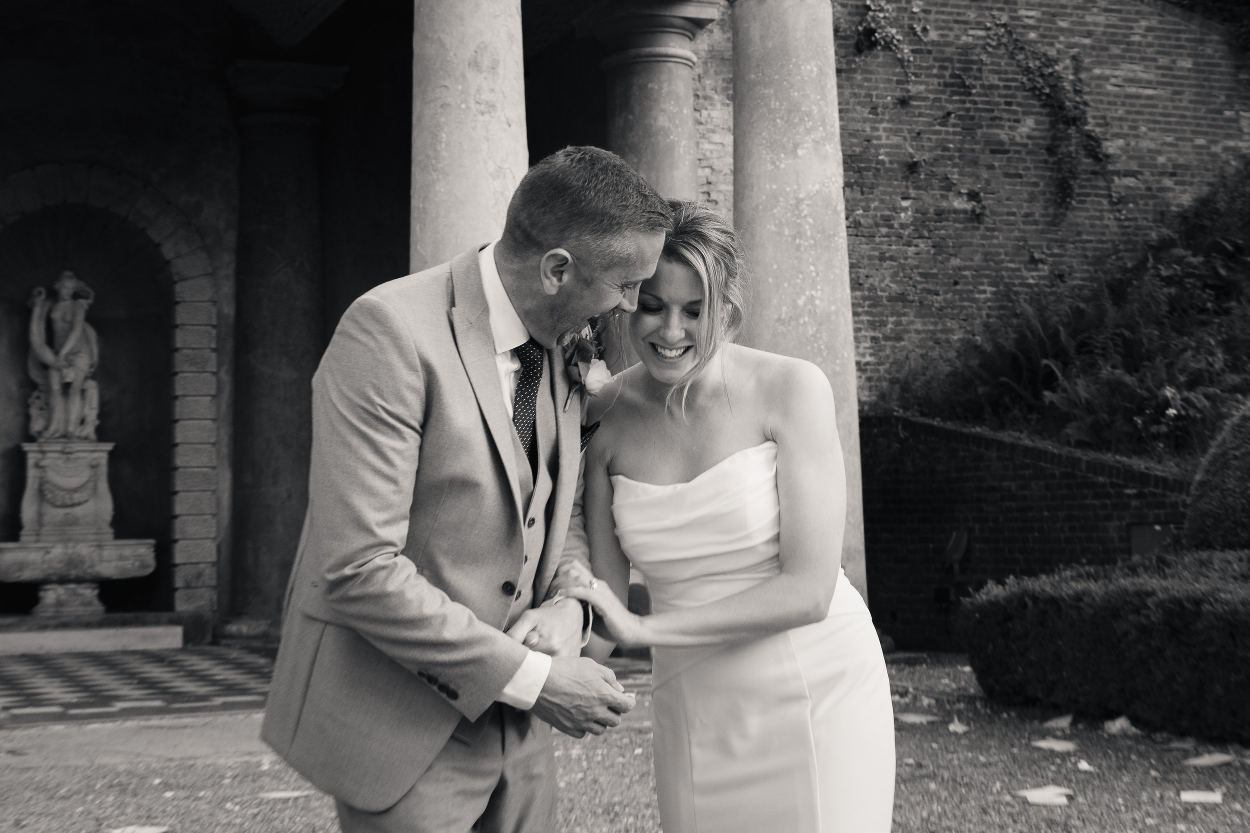 A black and white photo of a couple, possibly newlyweds, smiling and holding hands outside a historic brick building with columns and a statue in alcove, dressed in wedding attire.