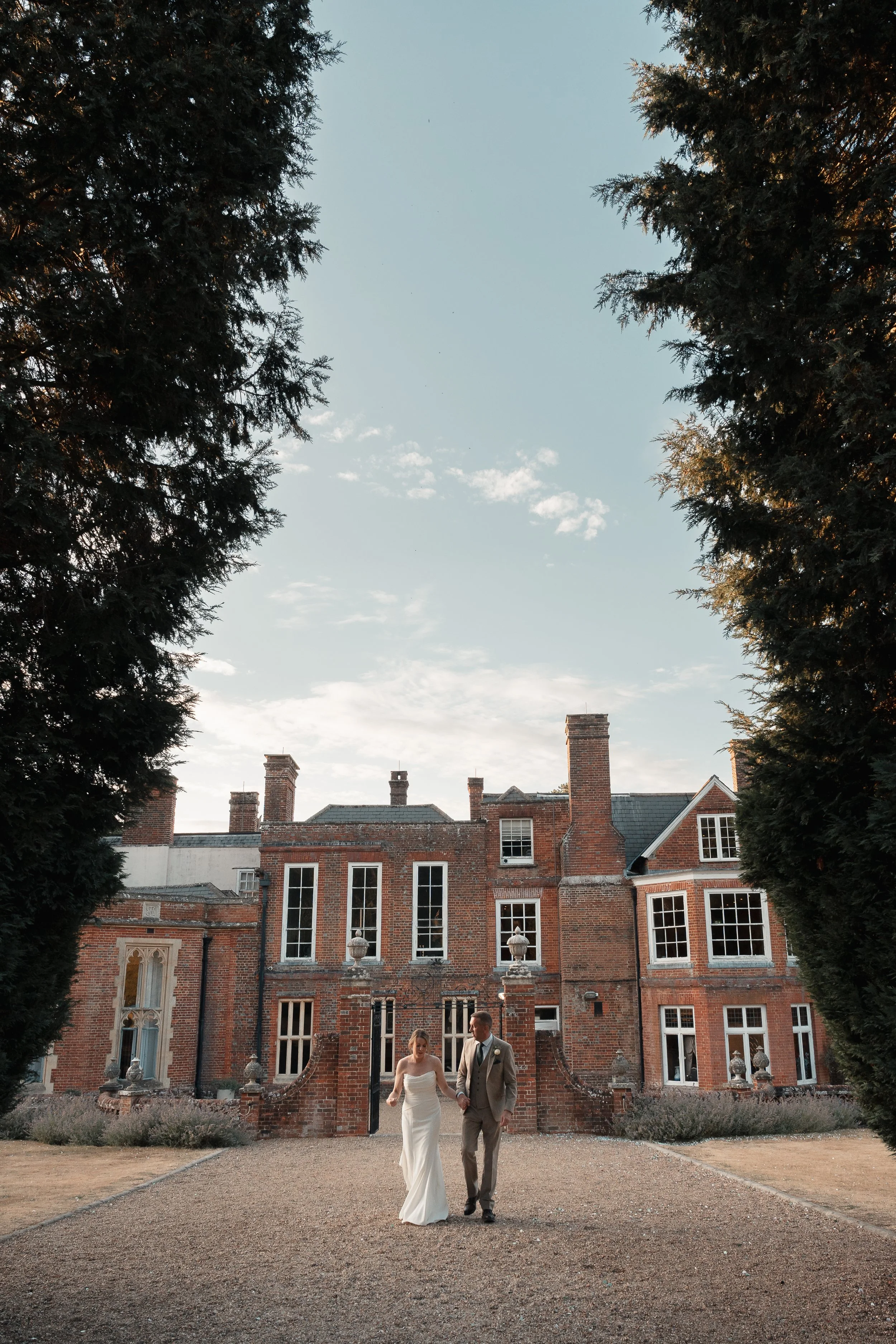 A bride and groom walking together outside a large red brick mansion with tall trees framing the scene, a gravel pathway, and a clear sky overhead.