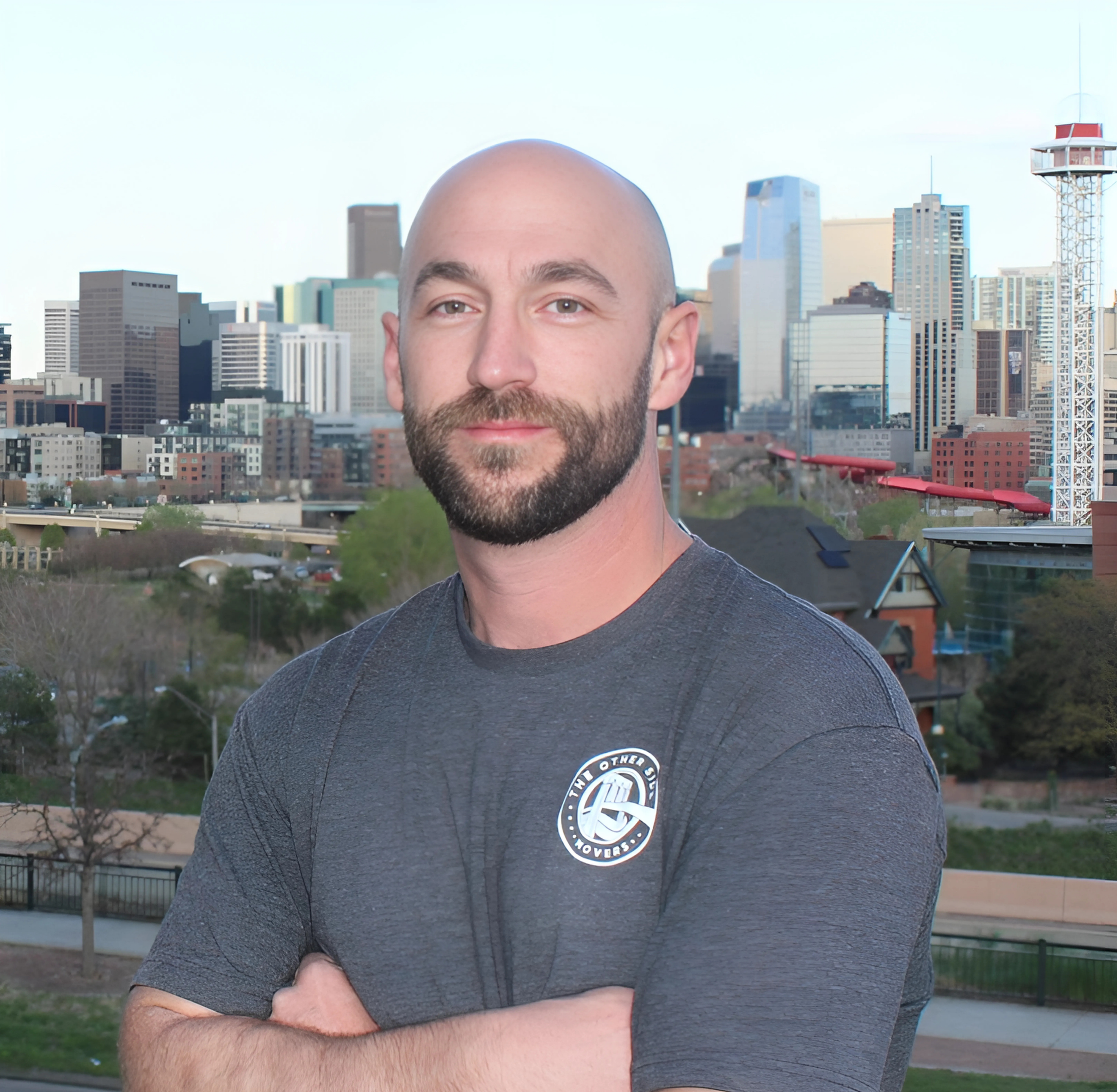 A man with a beard and shaved head standing outdoors with a city skyline in the background, wearing a gray T-shirt with a logo.