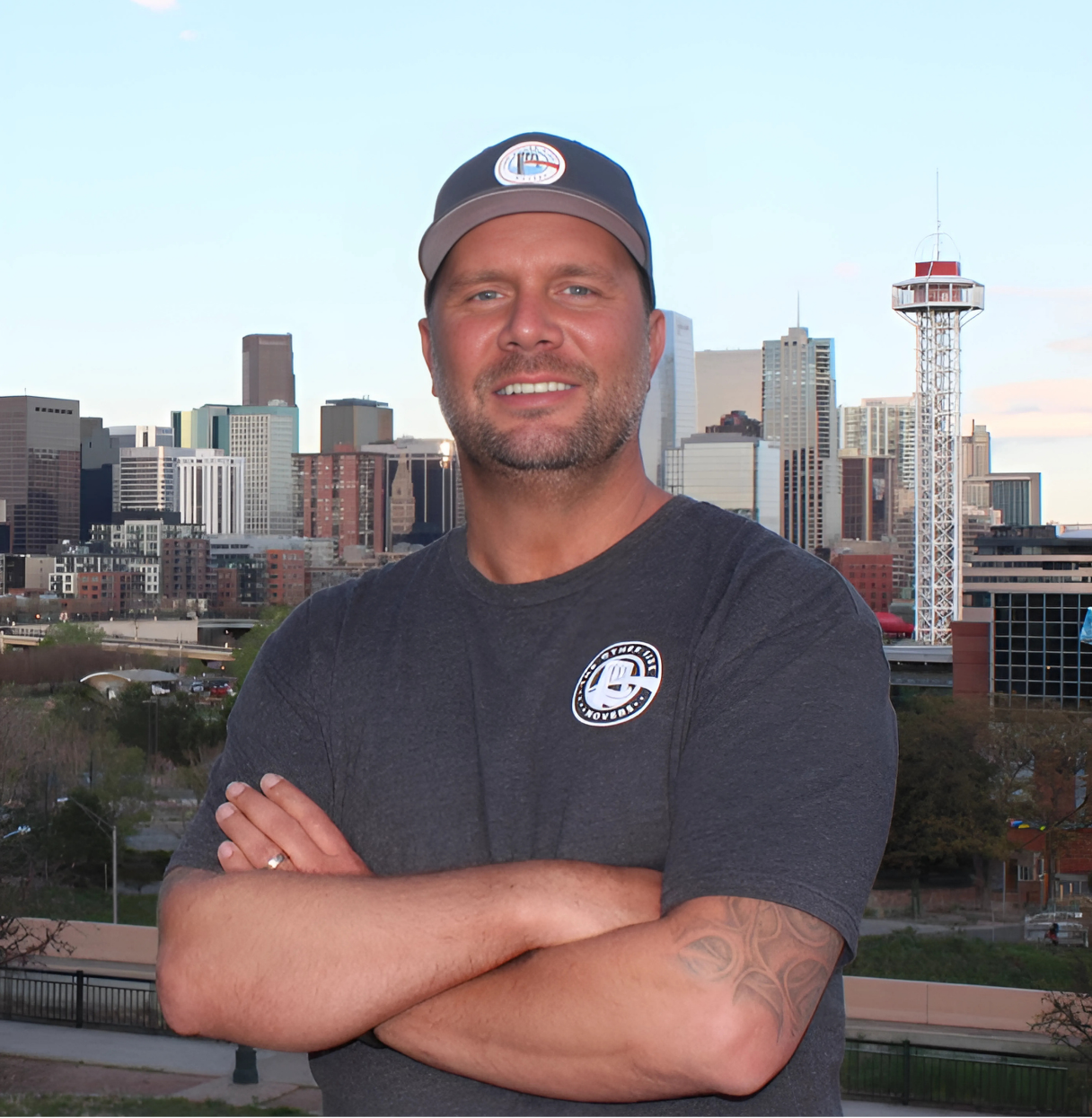 A man with crossed arms smiling in front of a city skyline with tall buildings and a tower during the daytime.