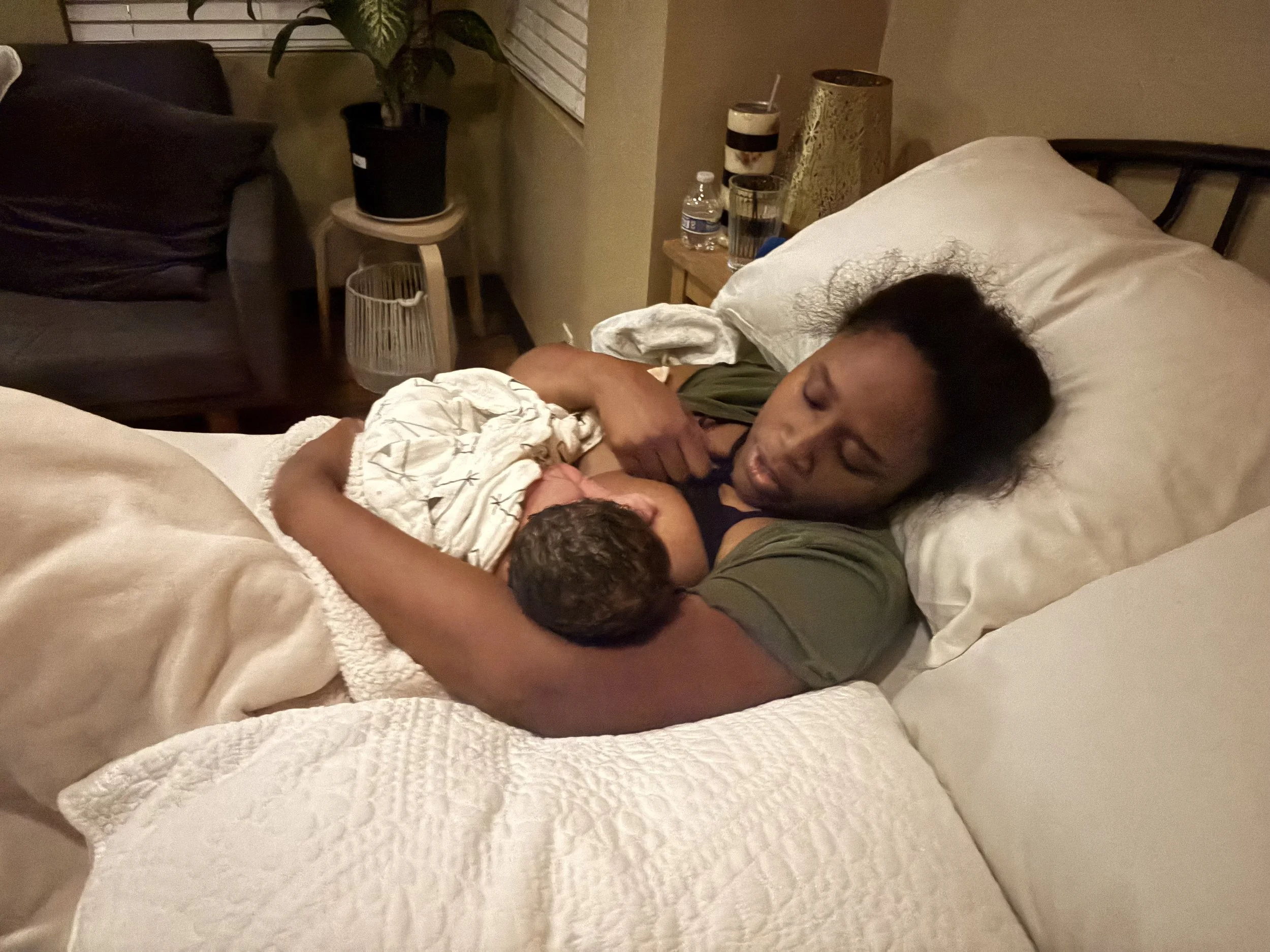 A woman lying in bed holding a small child, with a nightstand, water bottle, glass, and decorative items nearby.
