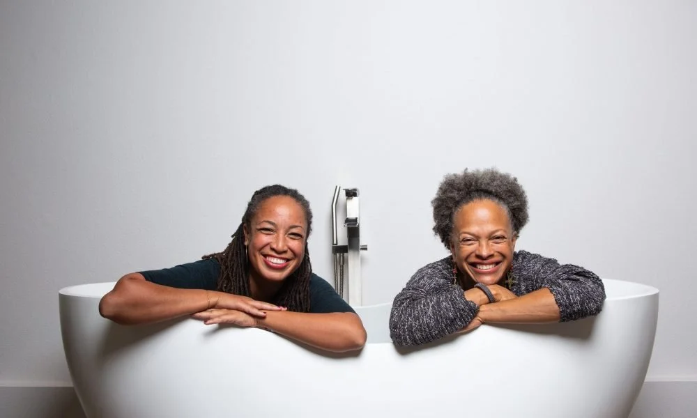 Los Angeles Birth Center founders and midwives Allegra Hill and Kimberly Durdin are smiling and relaxing in a white bathtub against a plain white wall.