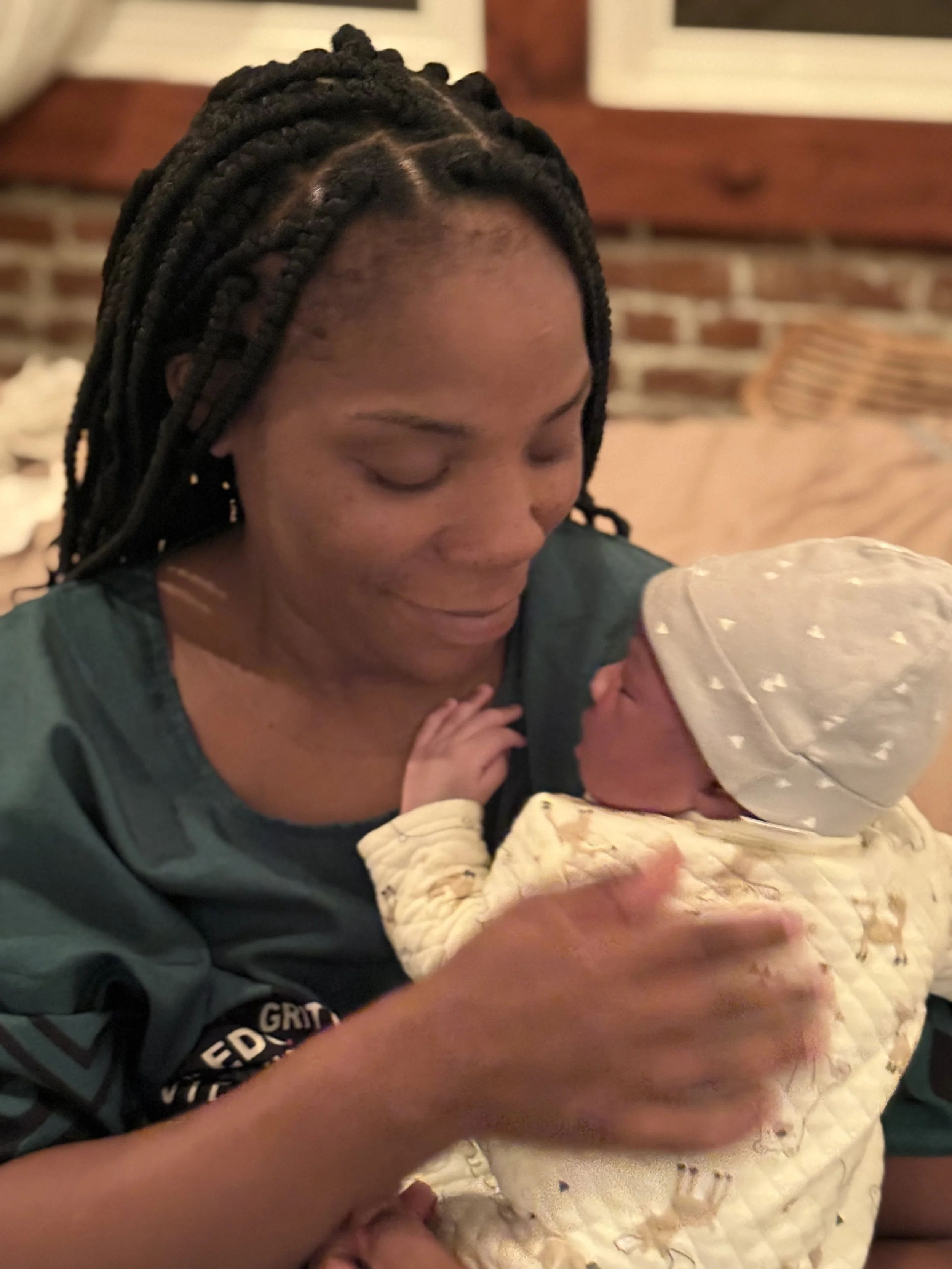 A woman with braided hair and closed eyes holds a baby wrapped in a yellow blanket and wearing a light-colored hat, in a cozy indoor setting.
