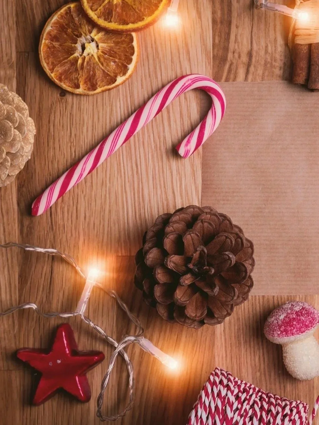 Holiday themed arrangement on a wooden table including a striped candy cane, dried orange slices, pine cone, red star-shaped decoration, string lights, and a mushroom-shaped candy.