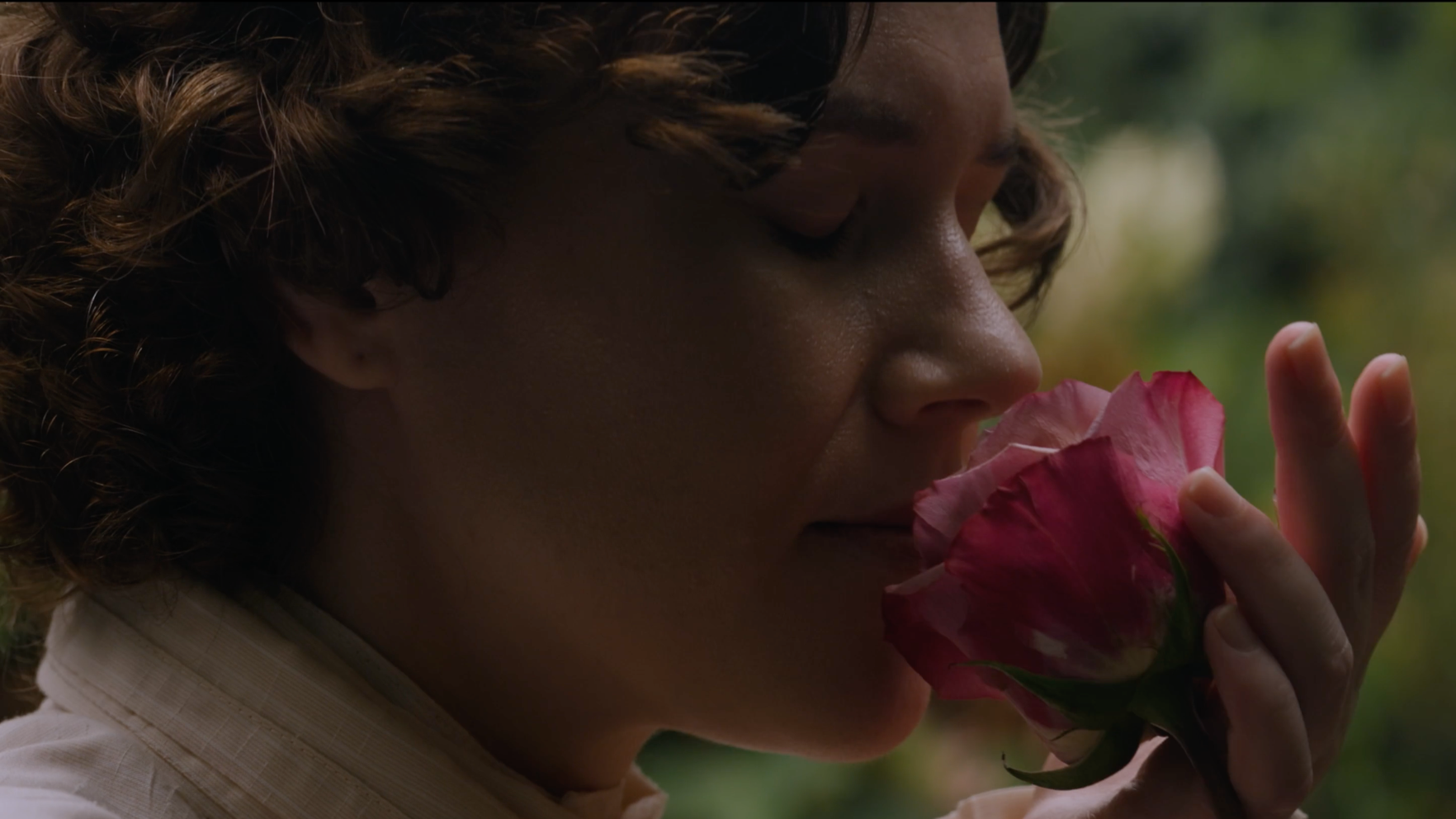 A woman with short curly hair and light skin holding a pink rose close to her nose with her eyes closed, outdoors with blurred green foliage in the background.