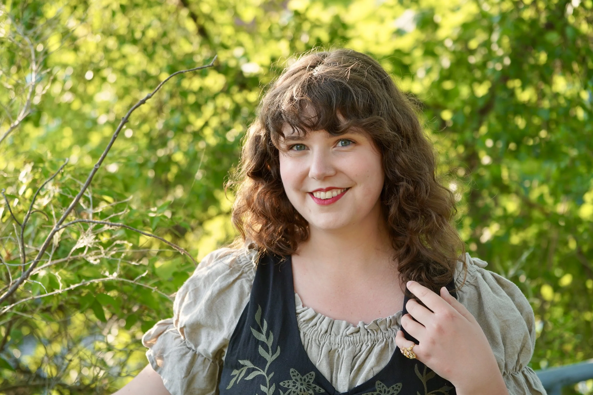 A headshot of Lauren Halász, the director. They are white and wearing a light green ruffled shirt with a dark green vest overtop. They have shoulder length dark brown curly hair with bangs and are wearing pink lipstick.