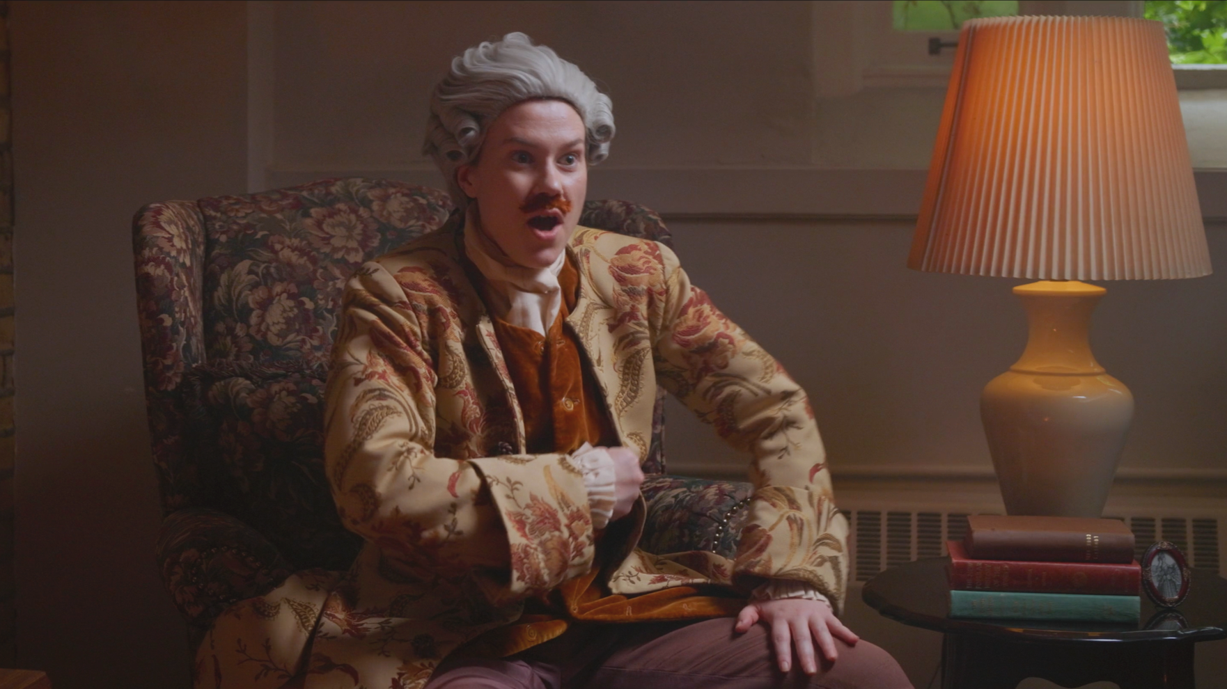 A person dressed in drag as a historical figure wearing an ornate jacket and grey wig. They're reacting with a silly suprised face.
