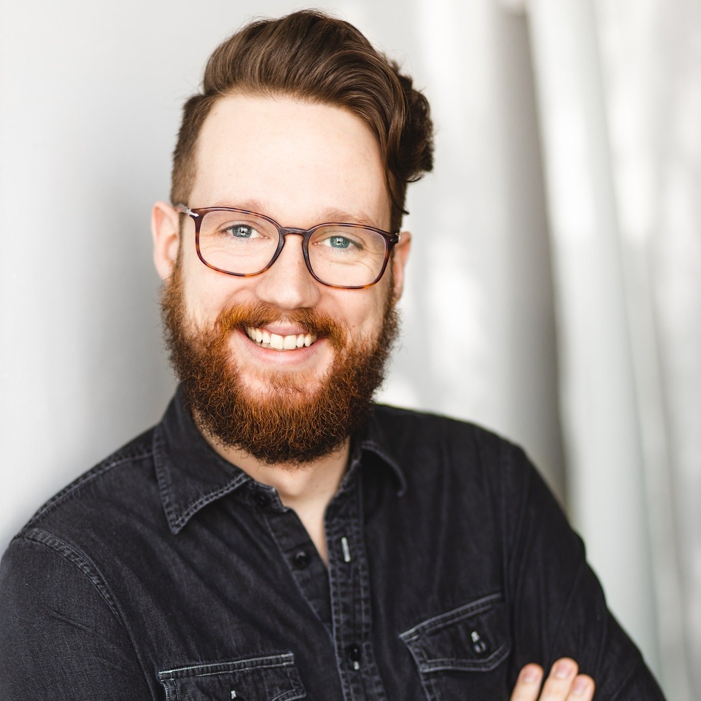 A headshot of Colin McMahon, the composer, a white person with short brown hair and beard with turtoise rectangle glasses. They’re wearing a dark denim button up.