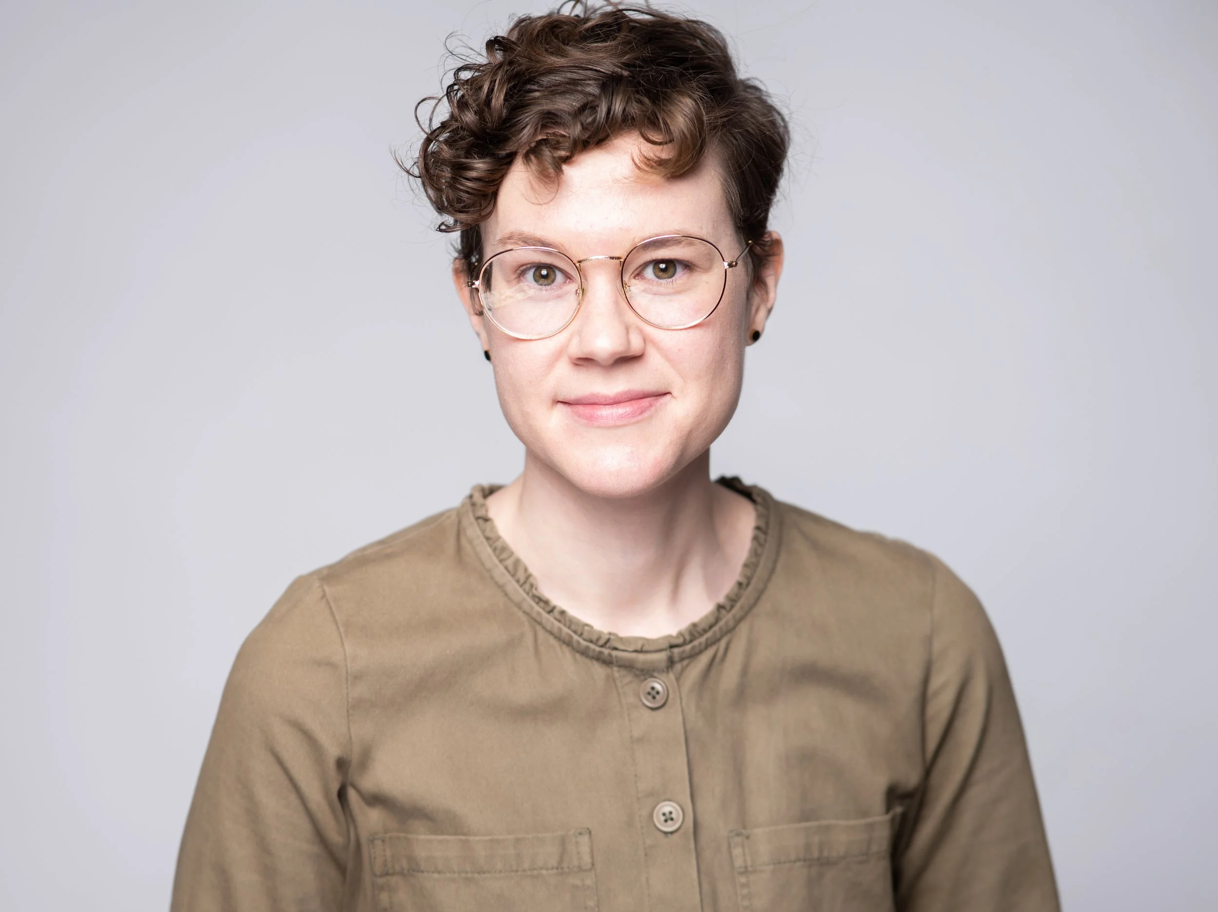 A headshot of Camille Rogers, the librettist and producer, wearing a light brown button up. They are white, have short curly brown hair and are wearing silver rimmed circular glasses.