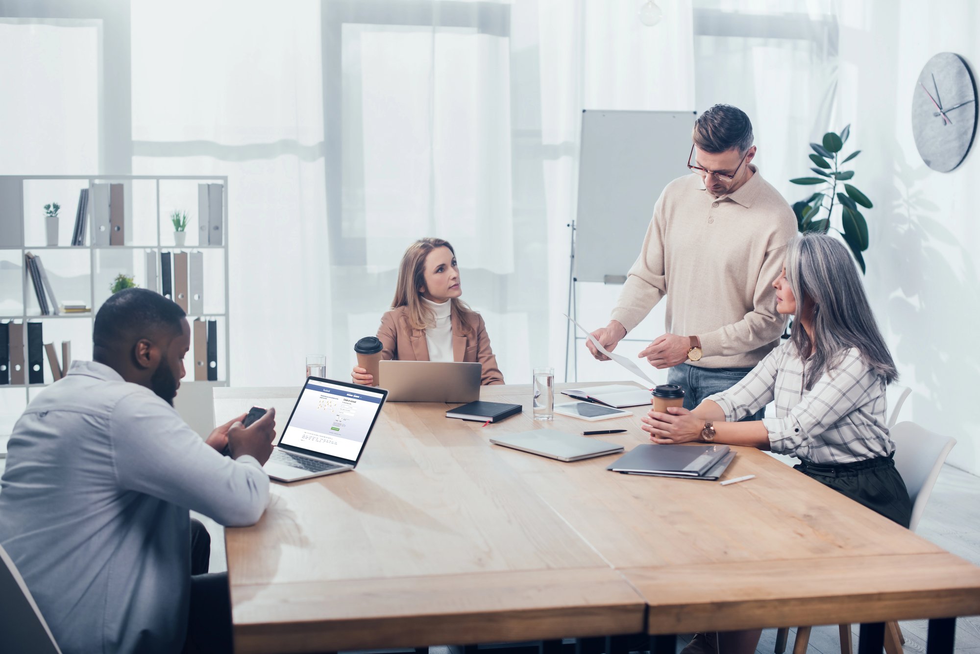 Woman with short hair sitting by a window, wearing a green scarf and eyeglasses on her head, looking thoughtfully to the side, with a laptop on the table.