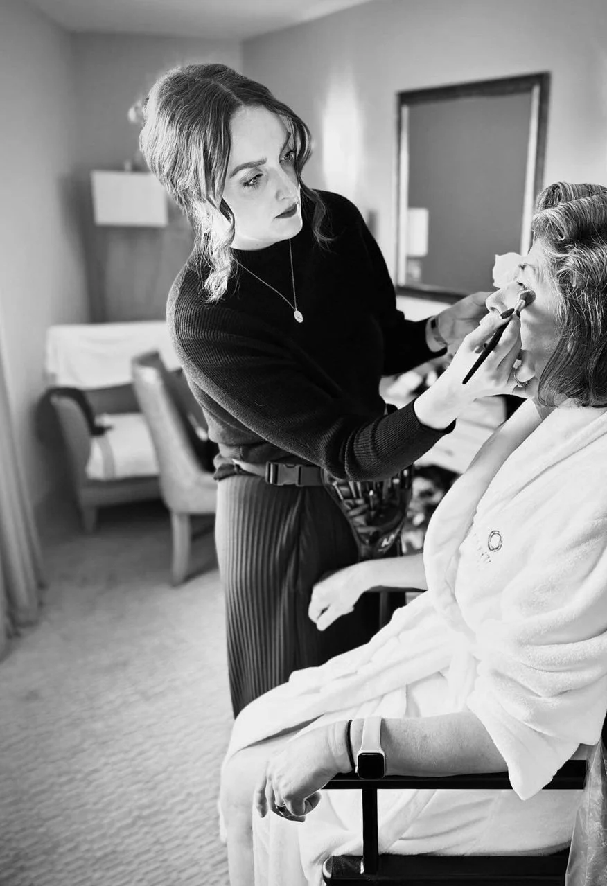 A makeup artist applies makeup to an older woman seated in a spa robe, in a cozy room with a bed and chair. Mother of the bride getting her makeup applied.