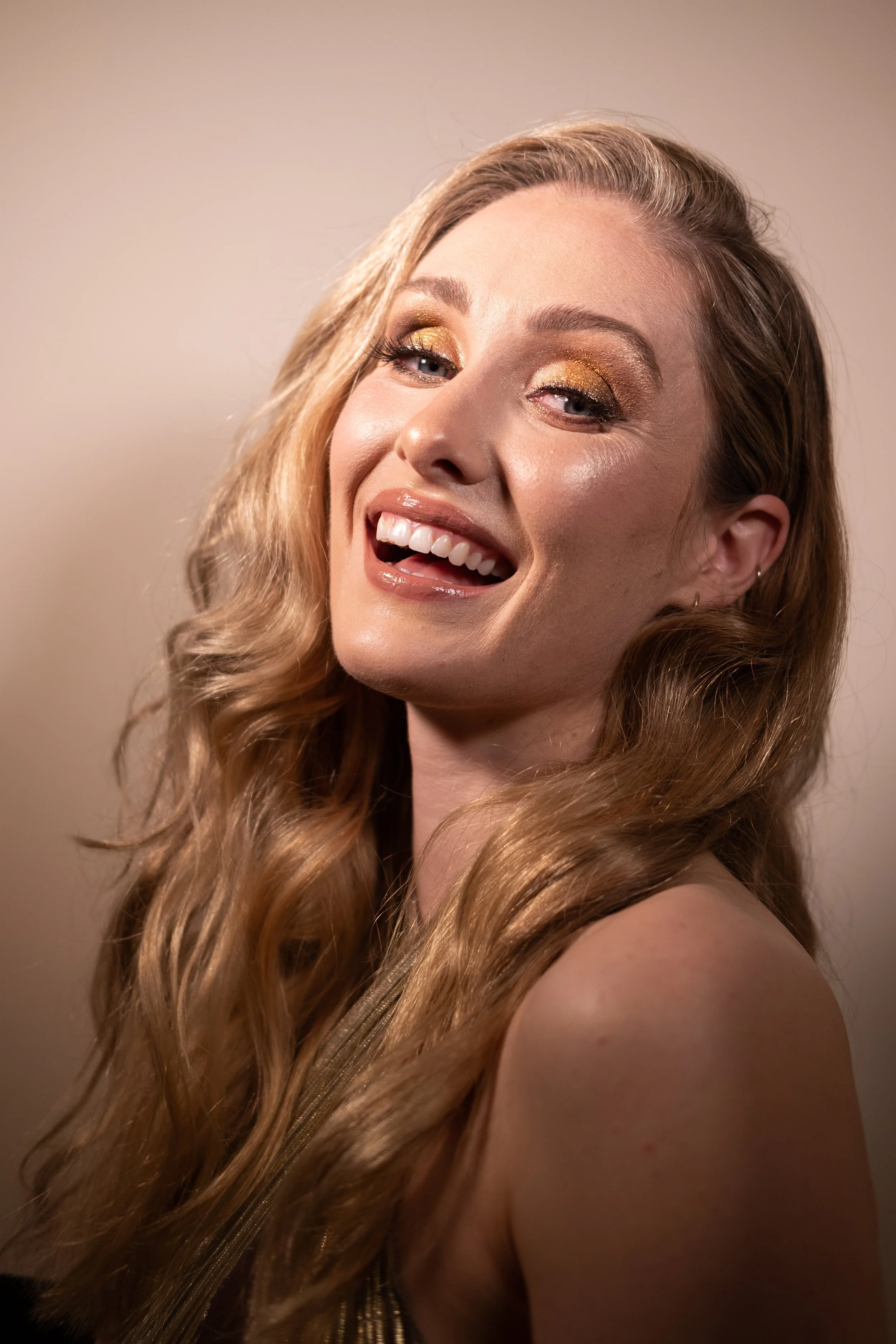 Close-up of a woman with long, curly blonde hair, gold eye makeup, and a wide smile showing her teeth, looking at the camera. Makeup and photography by @beautybybooms.