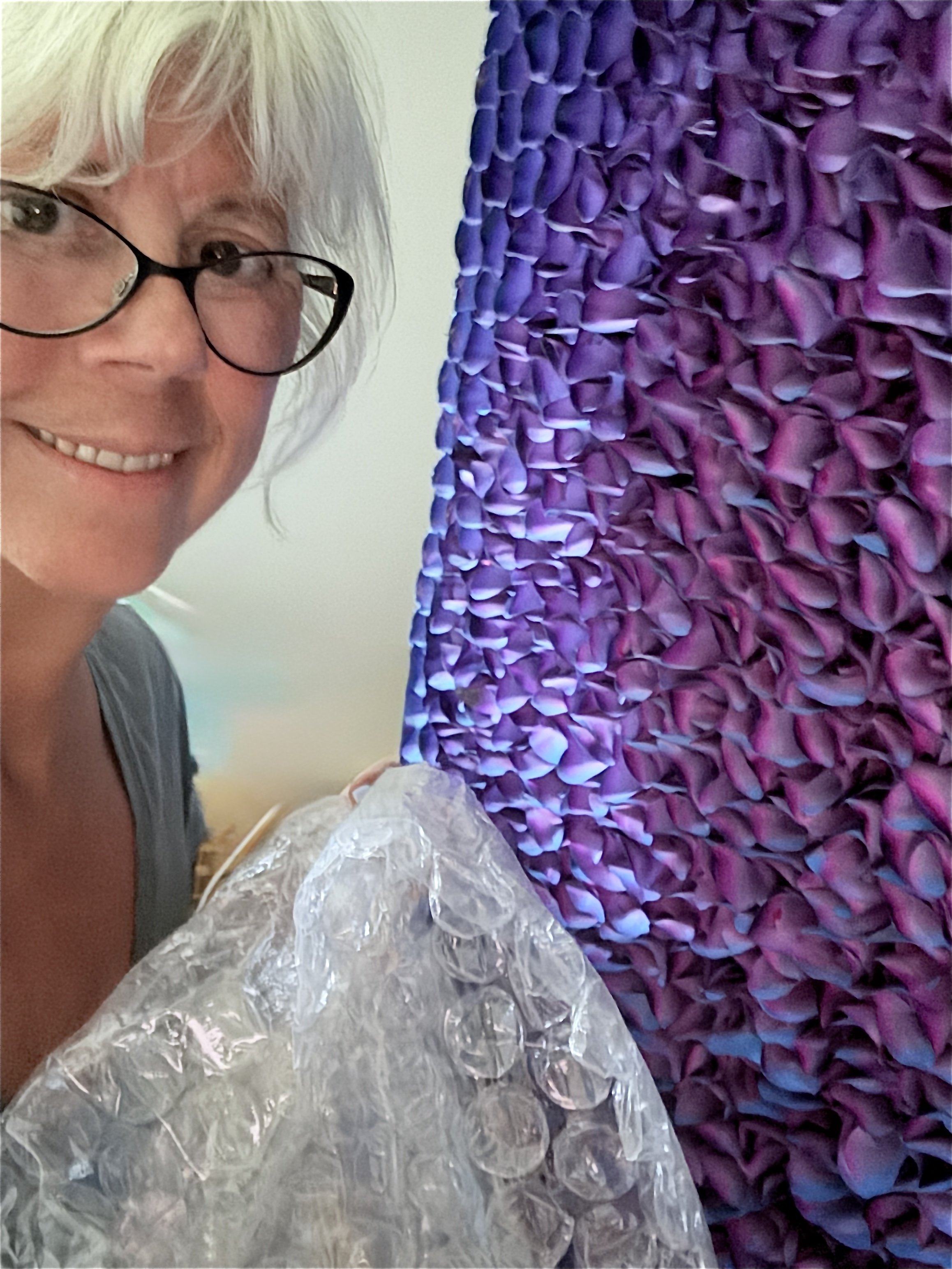 A woman with white hair and glasses smiling, holding a piece of clear plastic, stands next to a large purple textured surface.