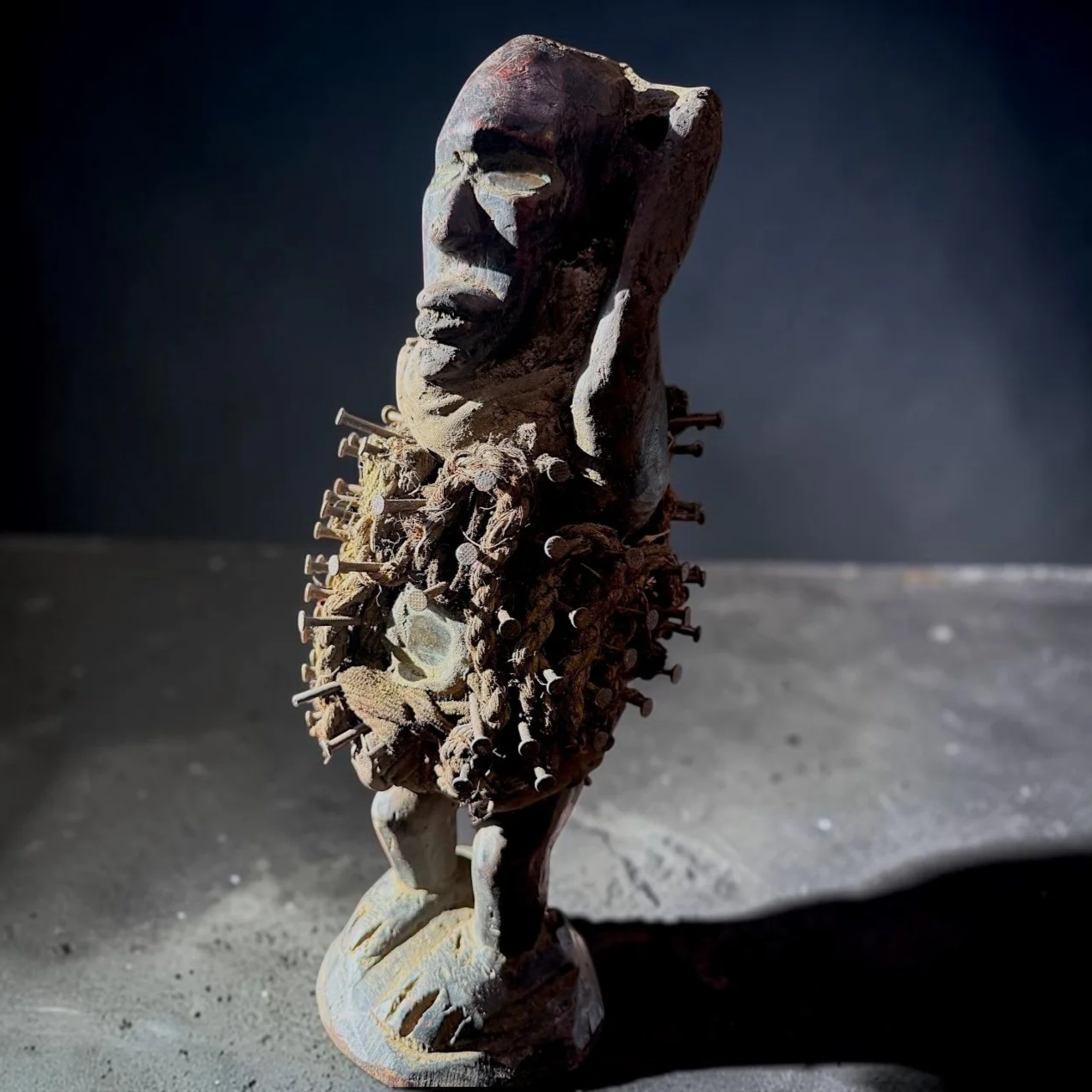 An art sculpture of a human face with nails and fabric around the neck, created from a stone or similar material, with a dark background and dramatic lighting.