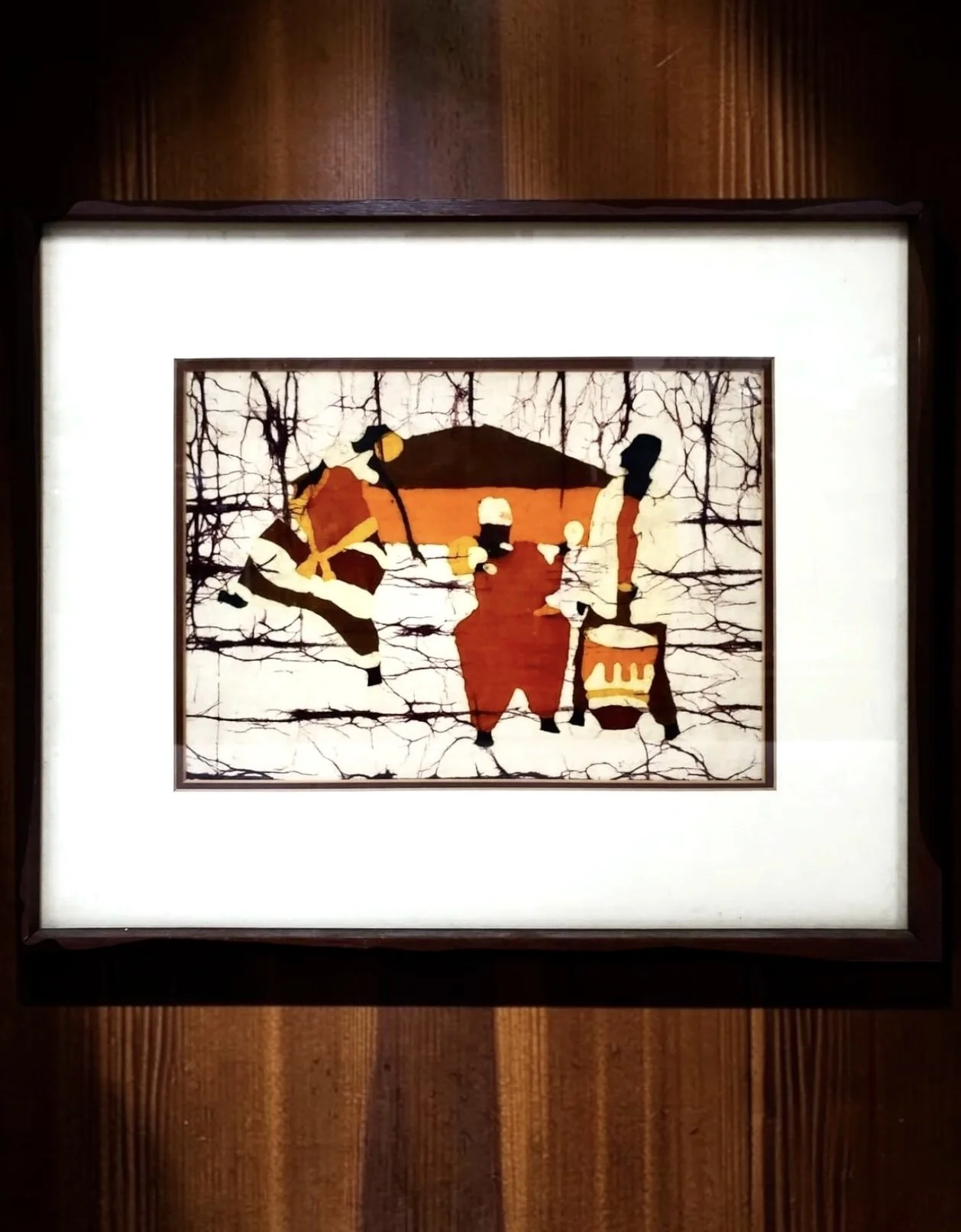 African Batik Figural Village Scene on Fabric
African batik on fabric, framed under glass, depicting three stylized figures in a rust, ochre, cream and black palette before a central hut and wooded ground, one figure carrying a drum, executed with th