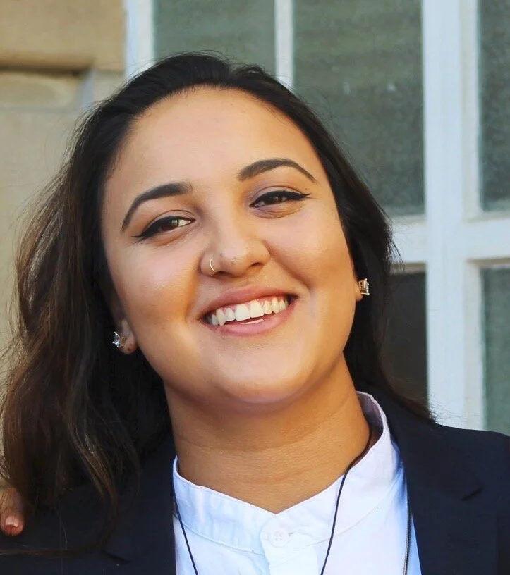A photo of a smiling Brown person in a white button up shirt and dark blazer