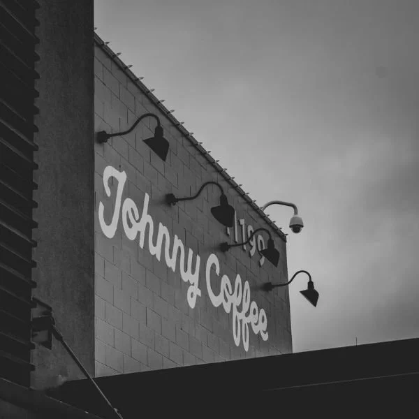 A white coffee mug with the text "Johnny Coffee" on it, placed on a light-colored surface with a shadow cast to the left.