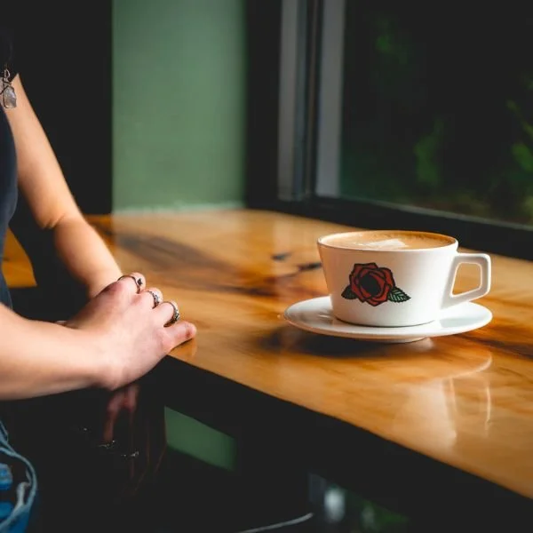 A clear plastic cup filled with iced coffee and cream, with swirling cream mixing into the coffee, placed on a wooden table near a window.