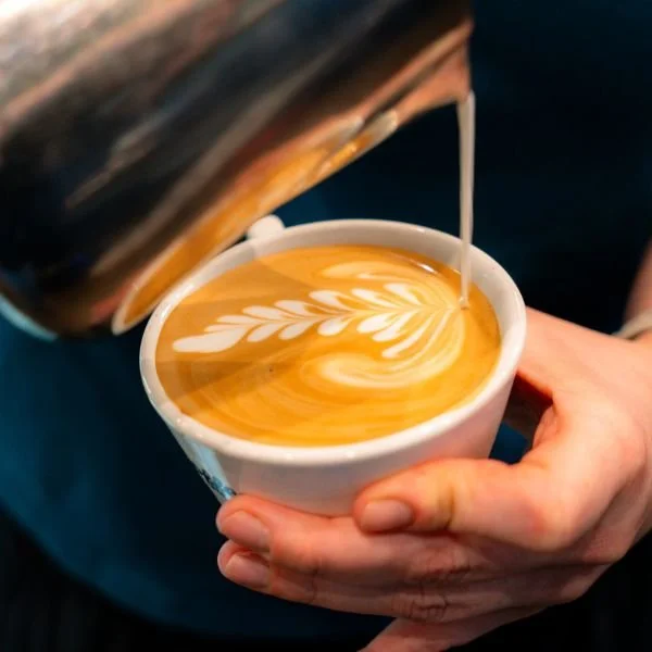 A barista pouring steamed milk into a cup of coffee, creating latte art with a heart design.