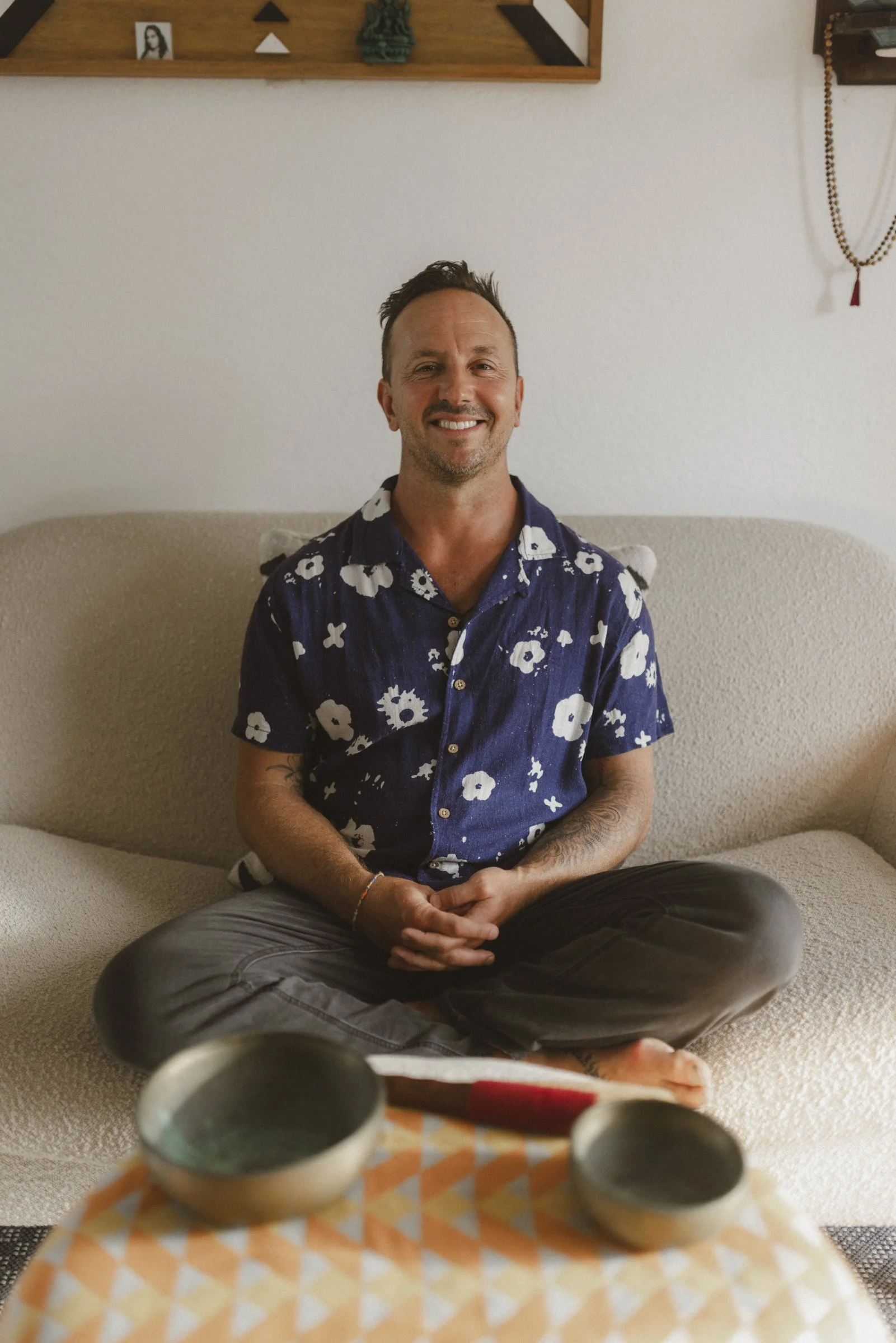 A man sitting cross-legged on a beige sofa smiling at the camera, wearing a blue floral shirt, with decorative bowls on a table in the foreground and wall decorations behind him.