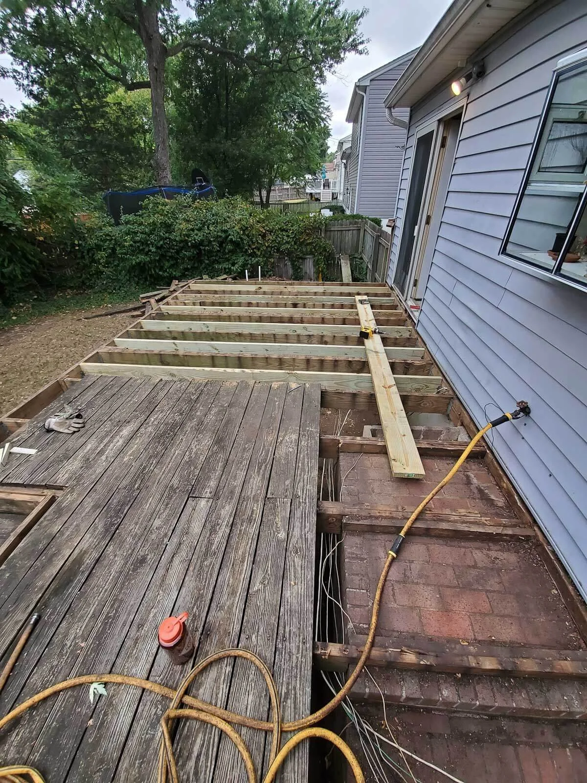 Backyard deck construction with wooden framework, electrical wiring, and power tools, next to a house with blue siding and a sliding glass door.