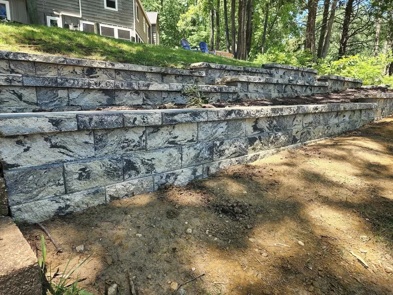 Stone retaining wall with multiple tiers, outdoors in a lush, green area, with a house in the background and blue outdoor chairs on the upper level.