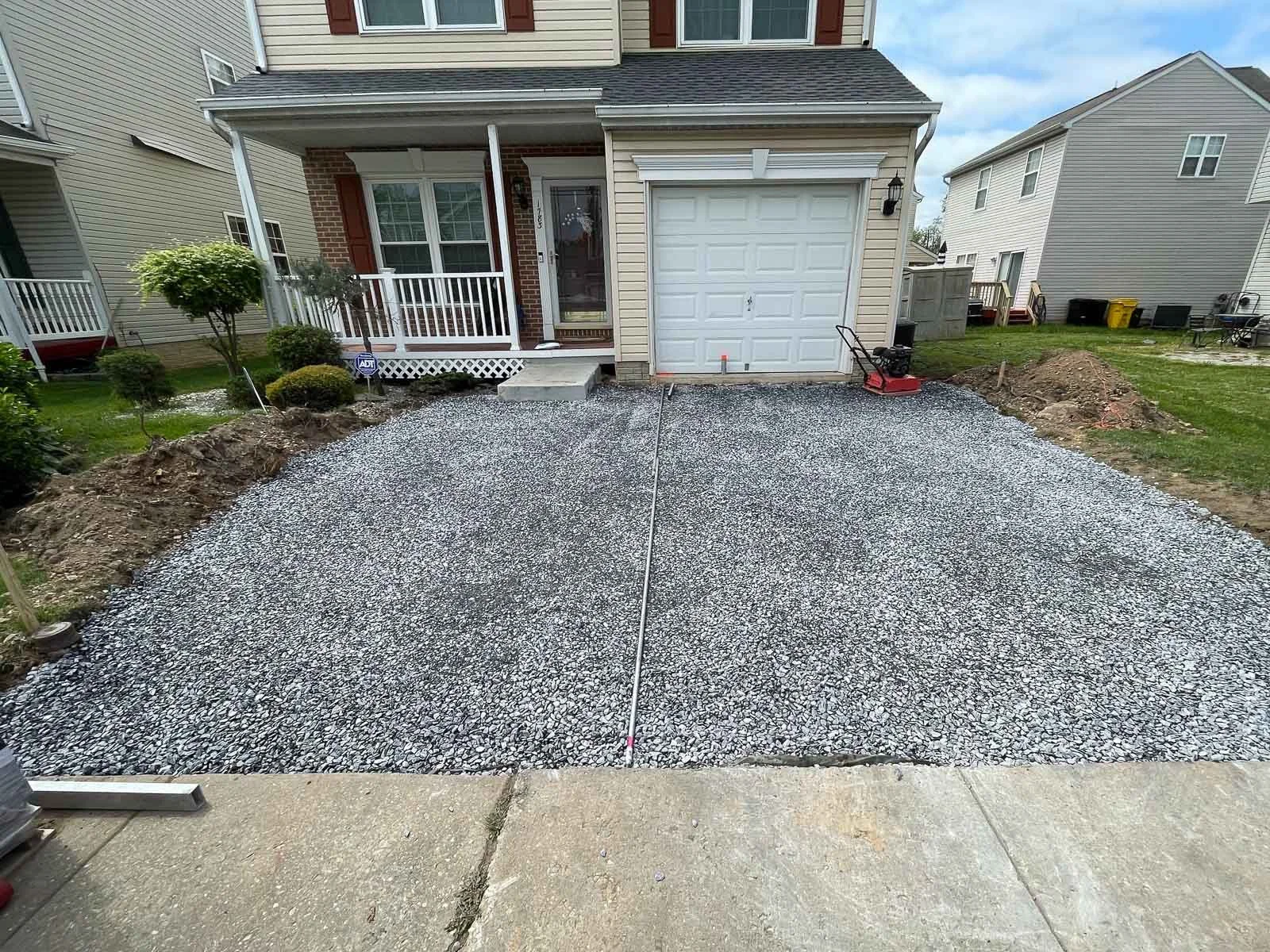A newly prepared gravel driveway in front of a house with a white garage door and front porch. The driveway is bordered by dirt on the sides for finishing, with some construction equipment and material visible.