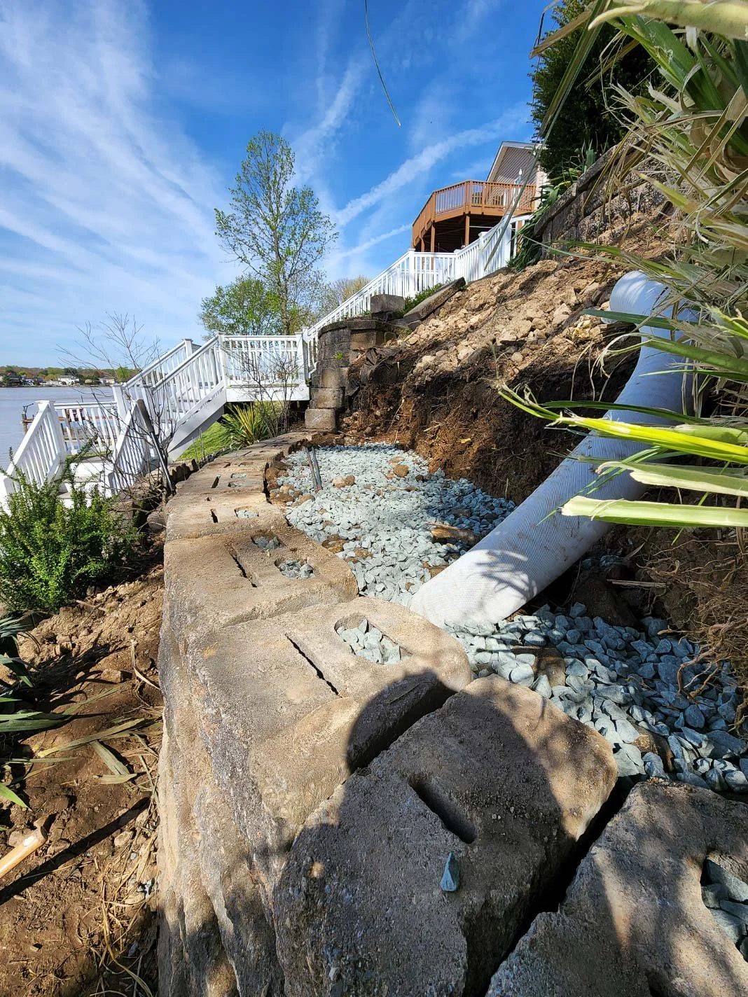 New retaining wall with large concrete blocks, a drainage pipe, and gravel foundation. White fences are along stairs leading to a house with a deck, with blue sky and trees in the background.