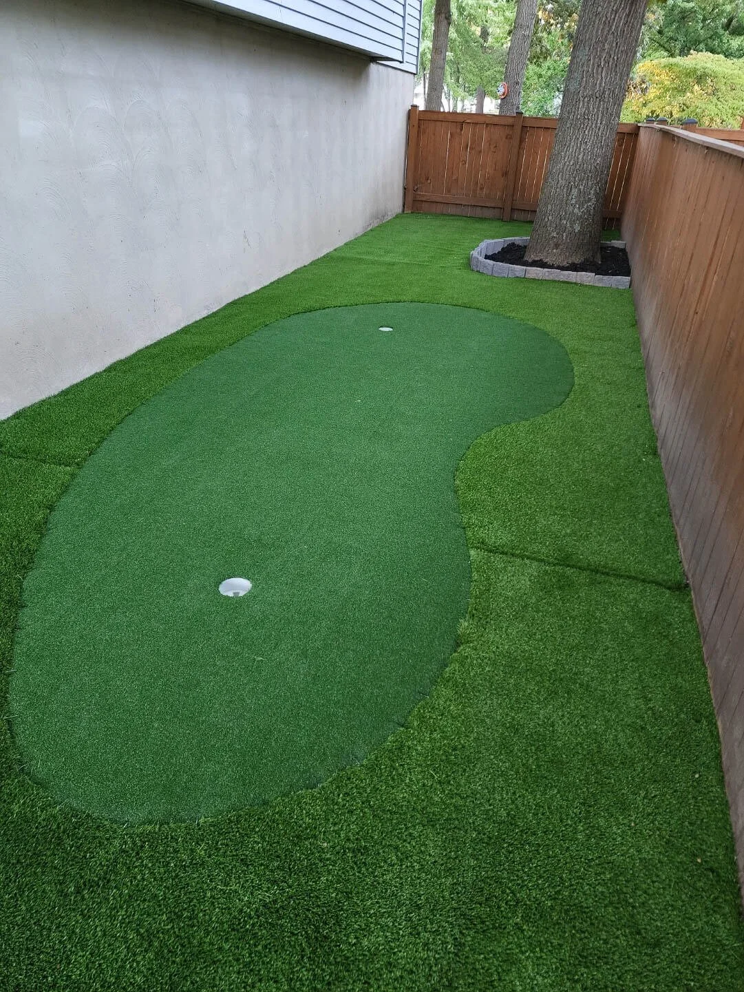 A small backyard renovated with artificial grass, two round golf hole cups, a tree surrounded by rocks, and wooden fencing.