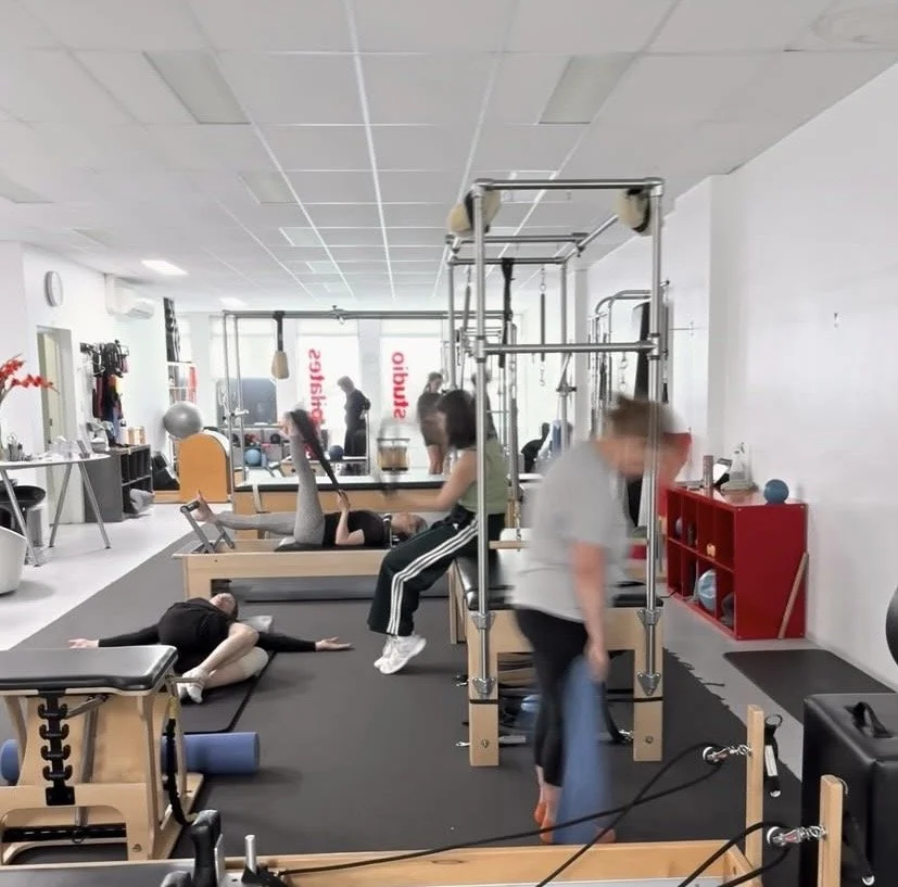 A busy fitness studio with people exercising on reformer machines and stretching on mats, with gym equipment and decorative items visible in the background.
