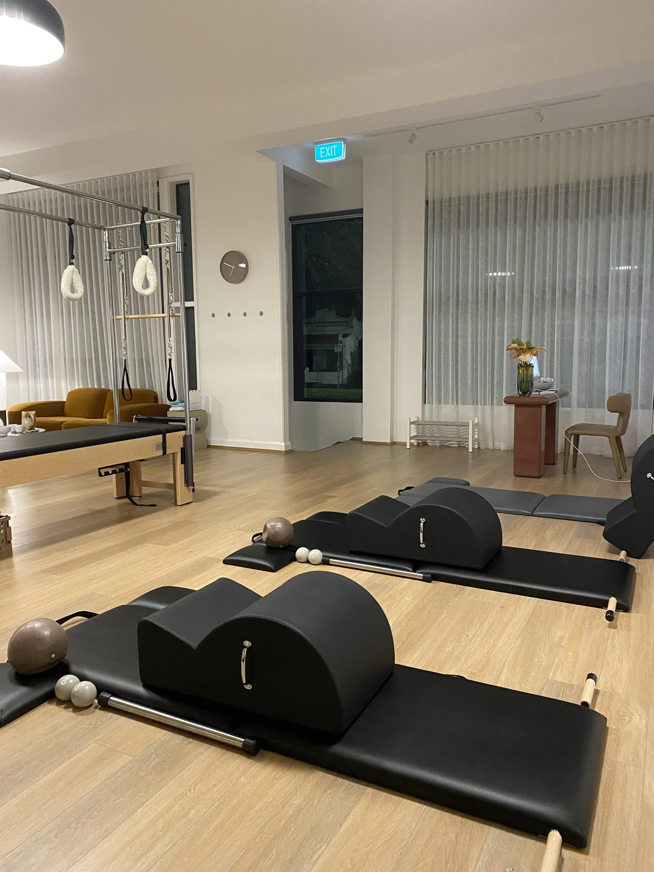 Fitness or physical therapy room with Pilates reformer machines, exercise balls, and a massage table. Contains a corner with a window, curtain, and a small table with a plant.