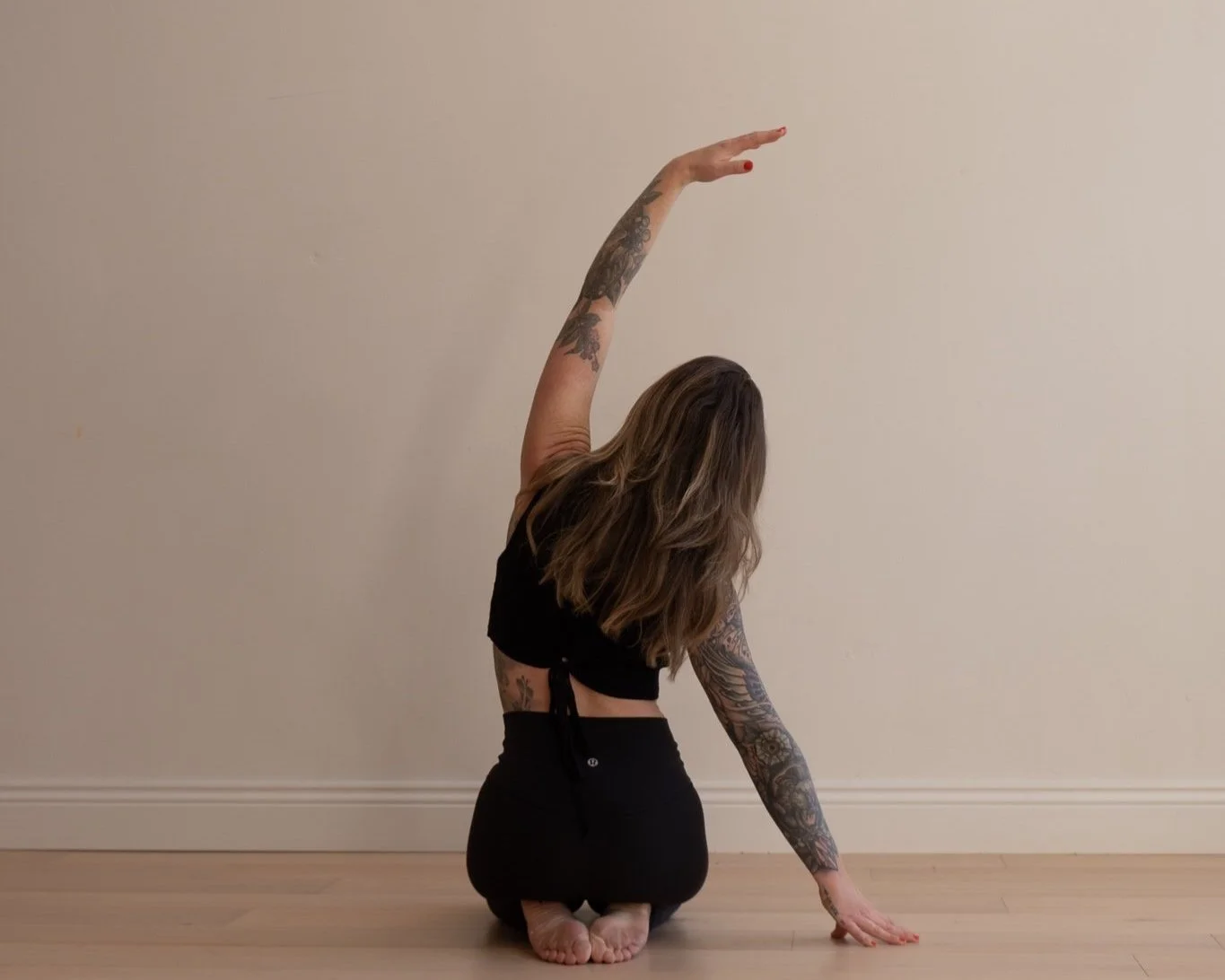 A woman with tattoos practicing yoga or stretching in a kneeling position against a plain wall.