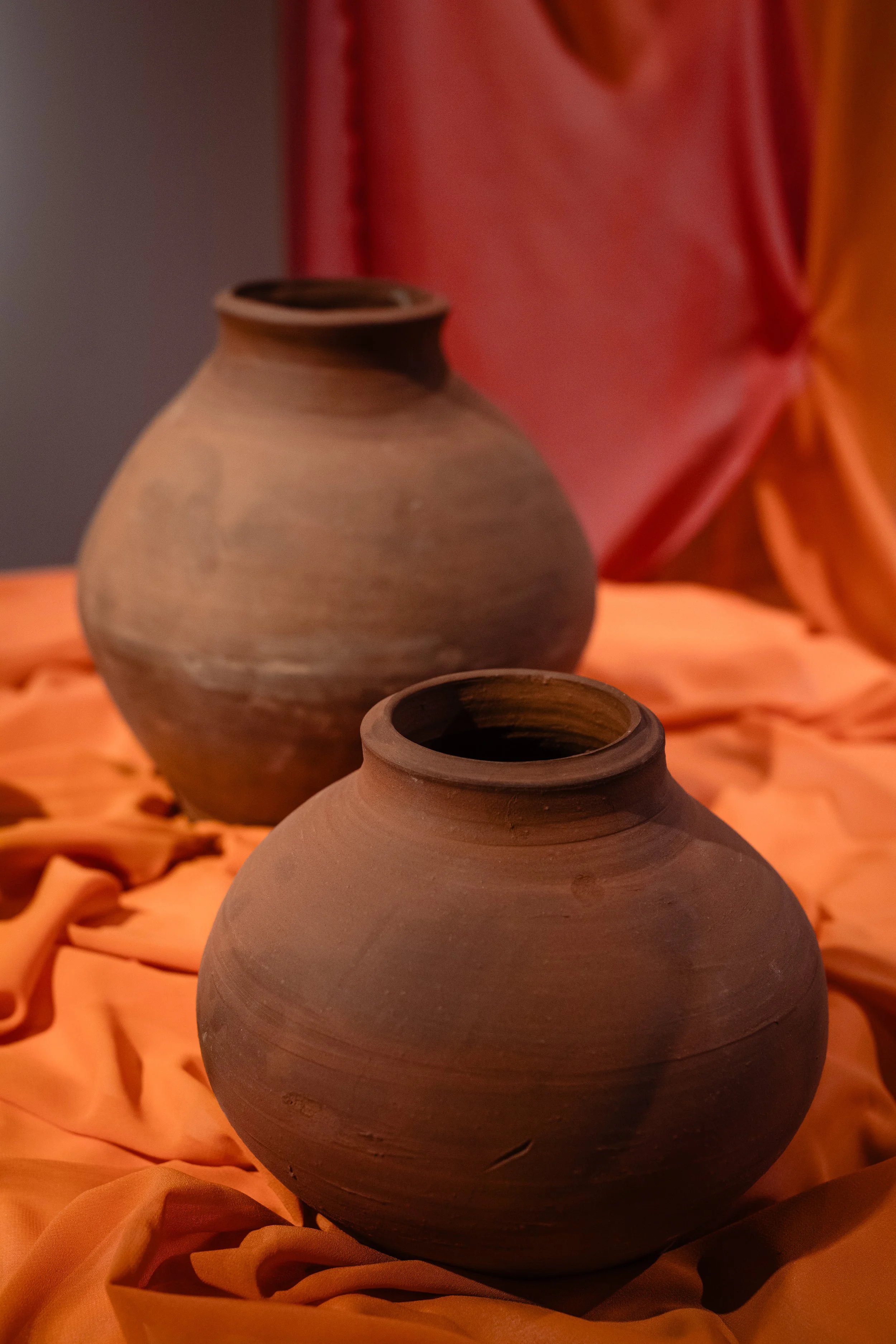 Two clay pots placed on orange fabric with red and orange draped background.