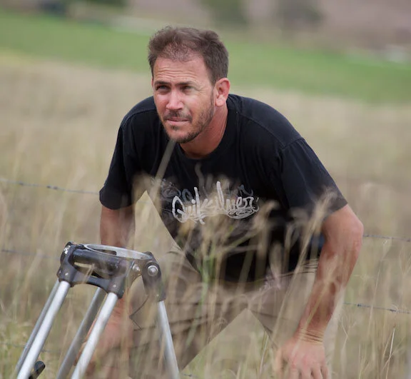 Chris Sun crouching on set in long grass beside a camera tripod