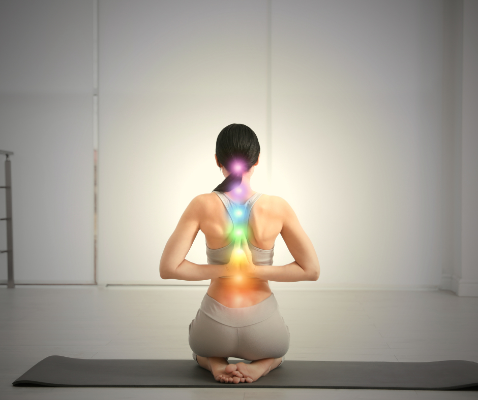 A woman practicing yoga indoors, seated on a mat, with colored chakras depicted along her spine.