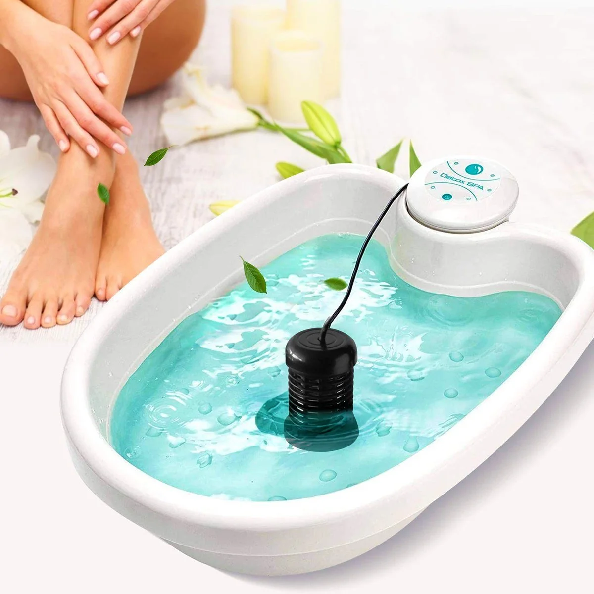 A foot detox machine in a white basin with blue water, surrounded by white flowers and candles, and a person receiving a foot massage in the background.