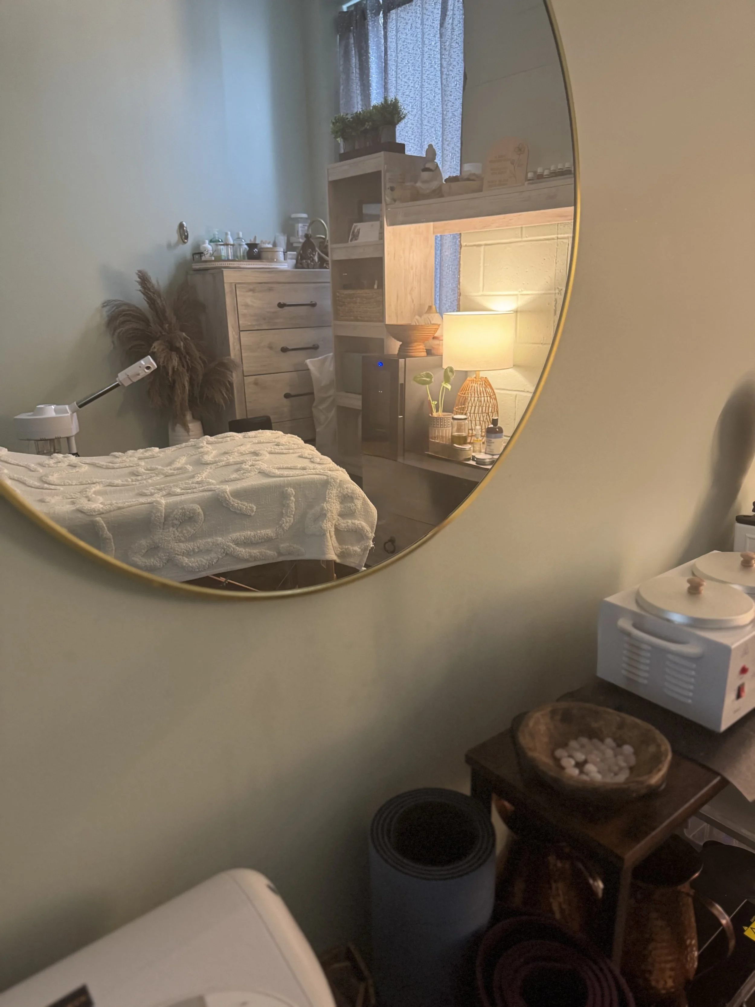 A reflection in a circular mirror shows a cozy room with a massage table covered in a white textured blanket, a wooden dresser, and a shelving unit with various items. A table lamp with a warm glow, a potted plant, and a window with curtains are also visible in the reflected scene. In the foreground, part of a white appliance and a small table with a bowl of cotton balls and a heat pad are seen.