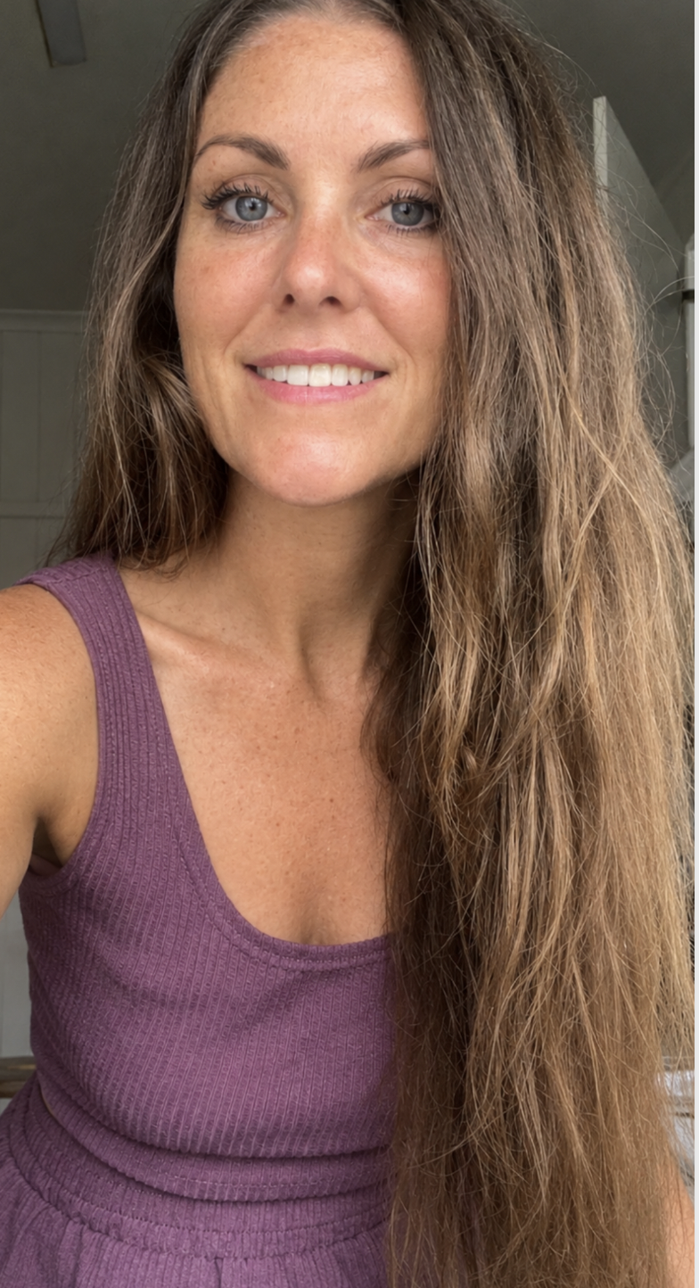 A woman with long, wavy light brown hair and blue eyes smiling, wearing a purple sleeveless top, indoors with neutral background.