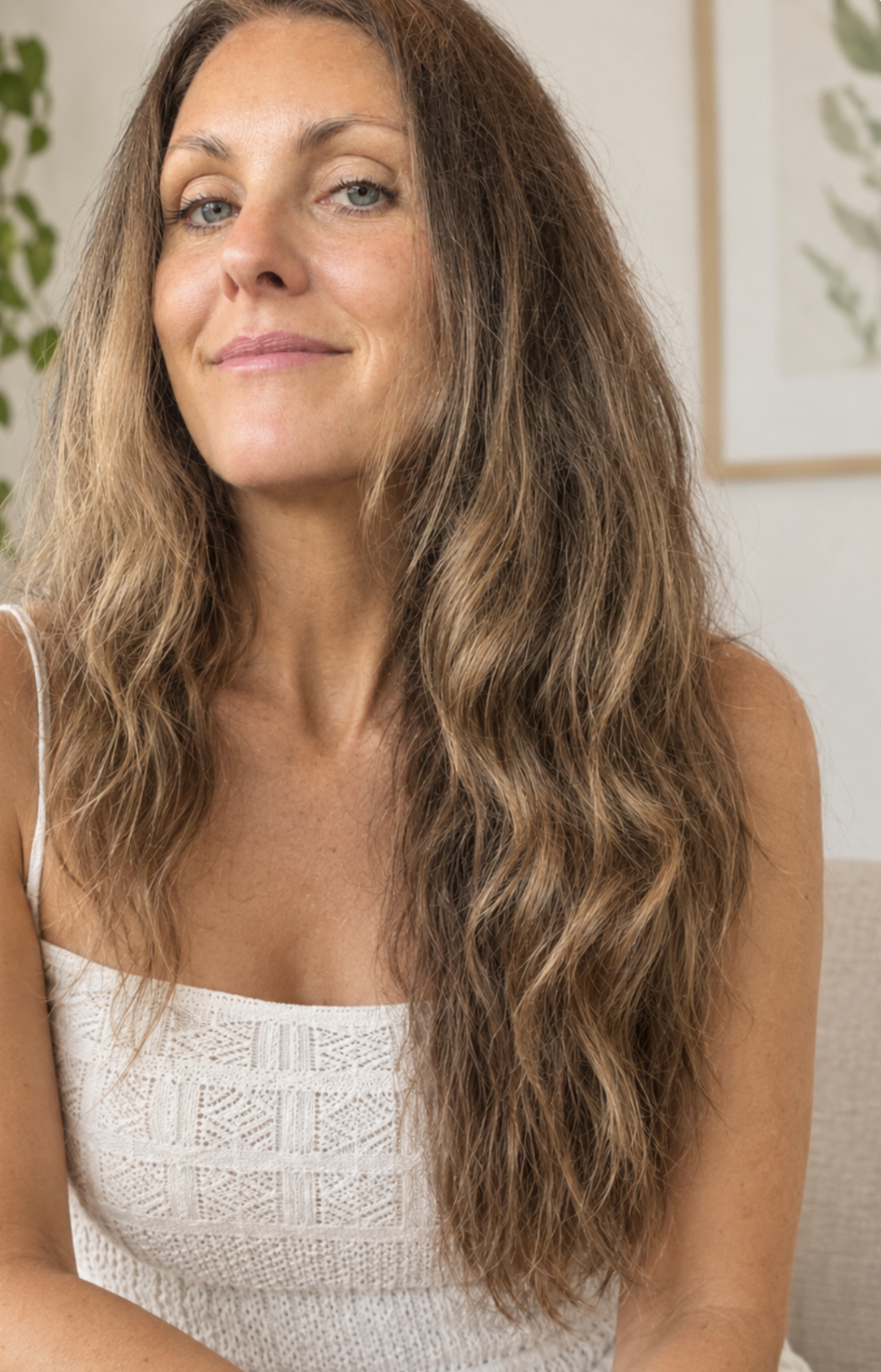 Close-up of a woman with long, wavy brown hair and light blue eyes, wearing a white sleeveless top, sitting indoors with houseplants and artwork in the background.