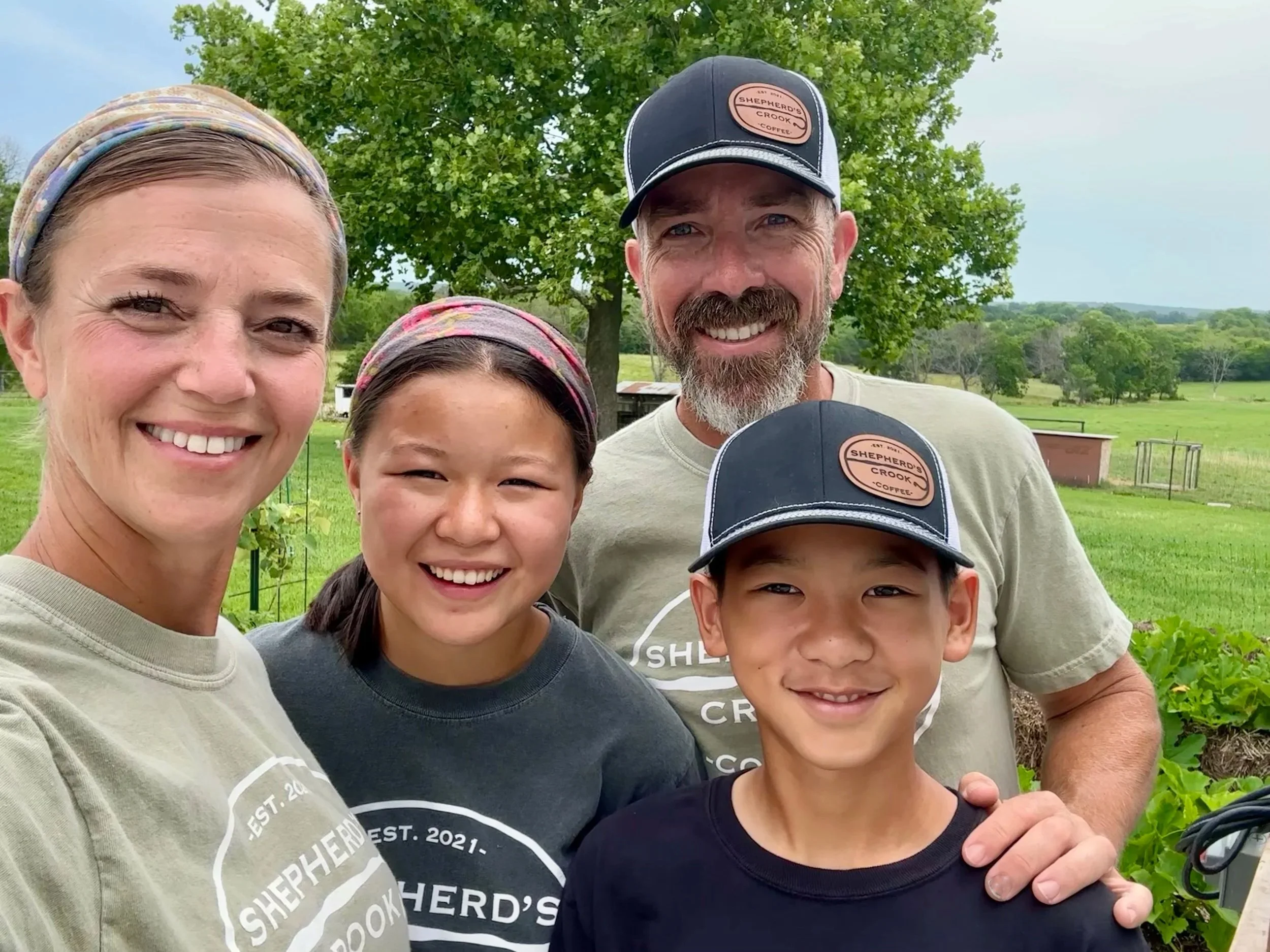 Family outdoors on a farm with green fields, trees, and a cloudy sky, smiling at the camera.